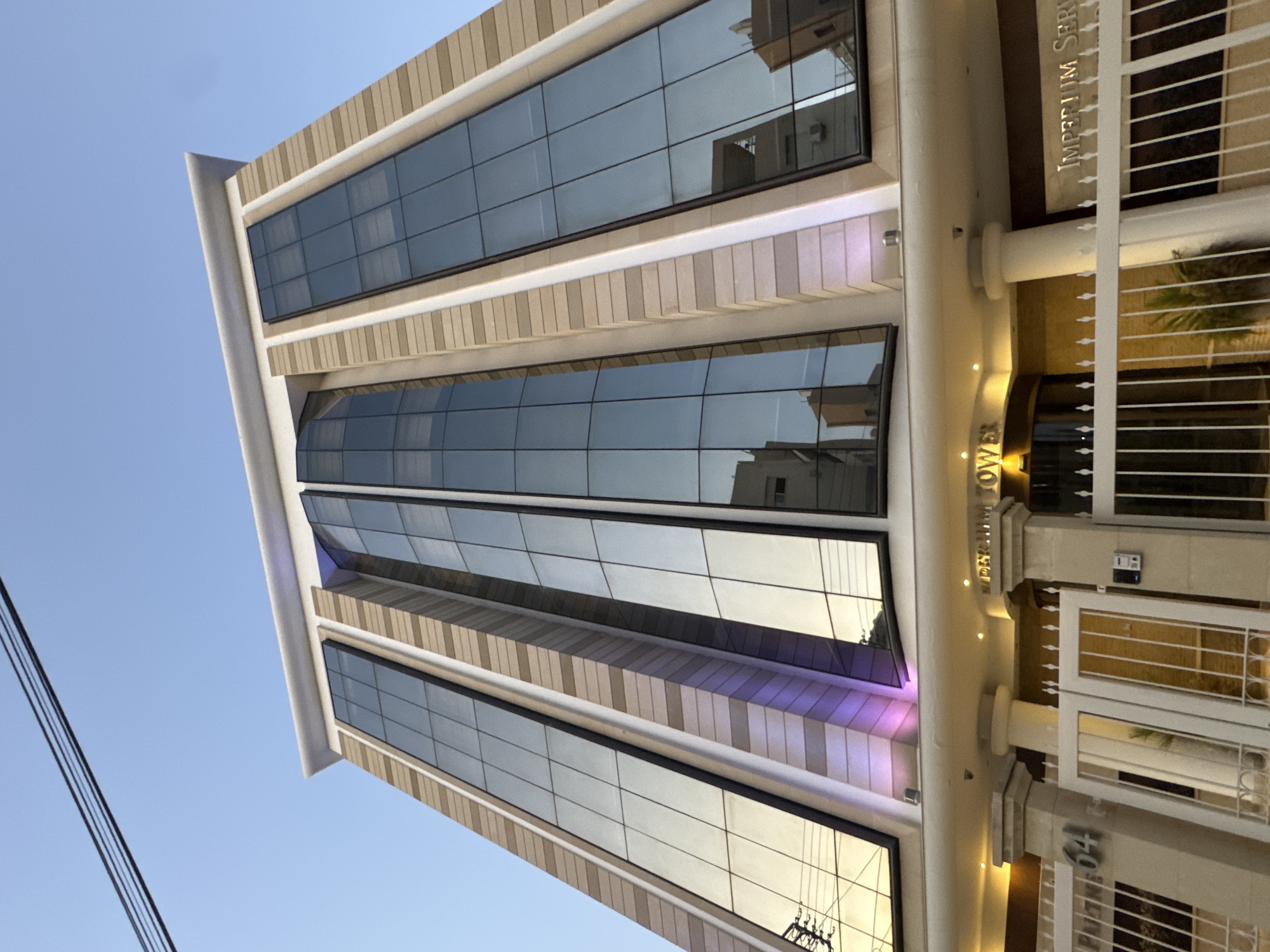 The image captures a modern, multi-story building with a facade of glass panels and beige-colored stone accents. The building appears to be an office or commercial space, with the name "IMPERIUM TOWER" visible in gold lettering above the entrance. The entrance features a revolving door and is flanked by pillars. A white metal fence with a gate is in the foreground, with the number "64" visible on one of the pillars. The sky is clear and blue, suggesting it is daytime. The building is located in Limassol, Cyprus, as indicated by the signage. The lighting on the building, particularly the purple illumination at the base of the glass panels, suggests it is either dusk or the lights have been activated. Reflections of other buildings and power lines are visible in the glass facade. There are no people visible in the image.
