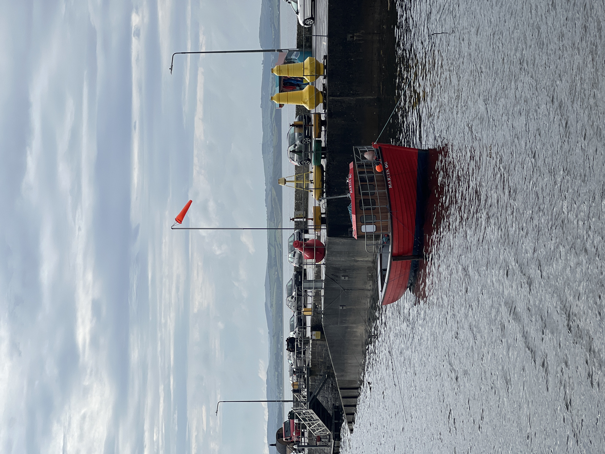 The image depicts a cloudy day at the harbor of Mountcharles, Ireland. A small red boat named "MARTIN OG" is moored in the foreground, its bow pointing towards the viewer. In the background, a concrete pier runs along the water, lined with several parked cars, including a red SUV, a silver sedan, a dark gray SUV, and a light gray hatchback. A metal walkway with stairs leads down to the water from the pier. There are also several buoys visible on the pier, two of which are large and yellow. A red windsock is visible on a pole to the left, indicating wind direction. The sea is calm with gentle ripples. Hills and a distant coastline are visible in the background.