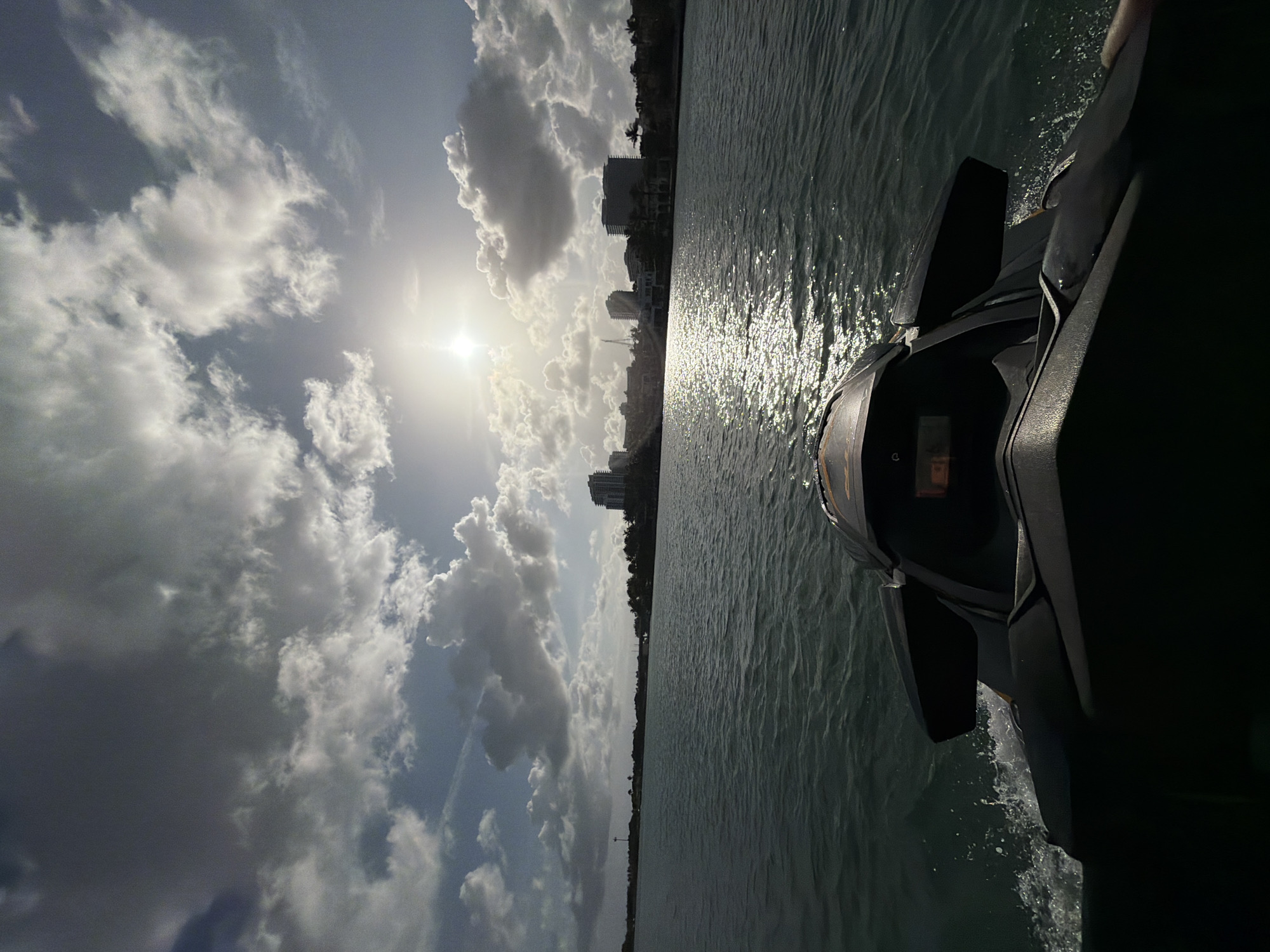 The image captures a scene from the water, likely taken from a watercraft, in Bal Harbour Village, United States of America. The sky is filled with a mix of clouds, some appearing dark and ominous, while others are bright white, reflecting the sunlight. The sun is positioned high in the sky, casting a glare. Below the sky, the water reflects the sunlight, creating a shimmering effect. The shoreline is visible in the distance, featuring buildings and vegetation. The watercraft is dark and partially obscures the view. The watercraft's wake is visible in the water.