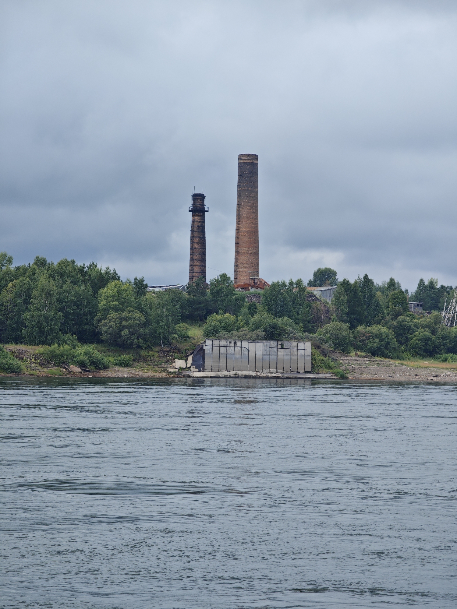 The image shows a wide river in the foreground, with ripples on its surface. In the background, on the riverbank, stands an industrial complex with two tall, brick chimneys. The chimneys are of different heights and appear aged. To the left of the chimneys, there are dense green trees and vegetation. In front of the trees, a concrete structure with vertical panels is visible, partially obscured by the greenery. The sky is overcast with grey clouds. This scene is located in Lesosibirsk, Russia.