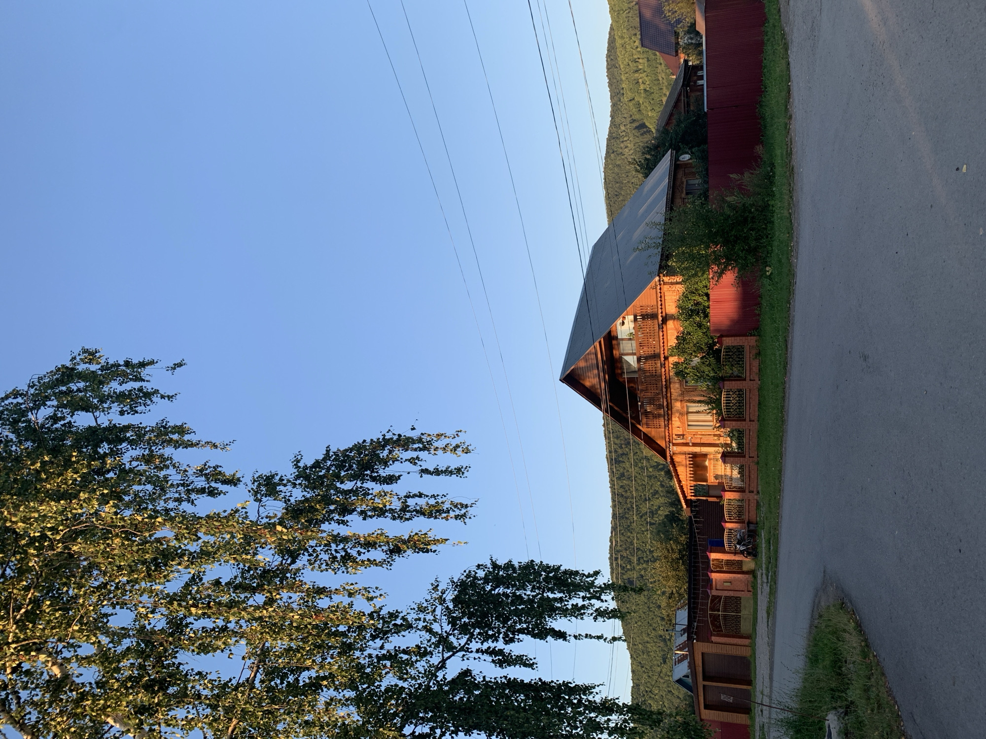 In Asha, Russia, a wooden house with an angled roof is situated on a street. The house is orange-brown and has balconies with railings. In front of the house is a red fence. To the left of the house is a smaller building with a garage. A paved road curves in front of the properties. Trees with green leaves hang from the upper left corner of the image. The sky is clear and blue, and a green, forested hill rises in the background.