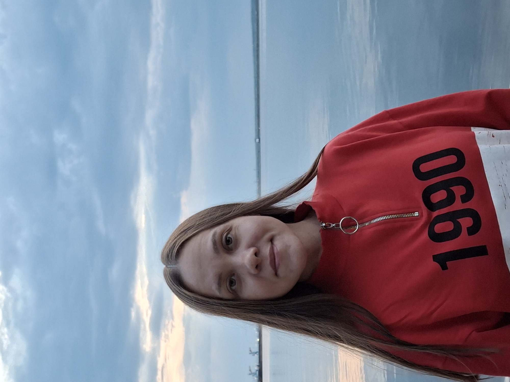 A woman is posing for a picture on a pier. The pier overlooks a body of water. Behind the water is a bridge. In the background, on the right side, are small cranes and docks. The sky is blue and has white clouds. The woman is wearing a red sweatshirt that has the number 1990 in black. The location is Chaikovsky, Russia.