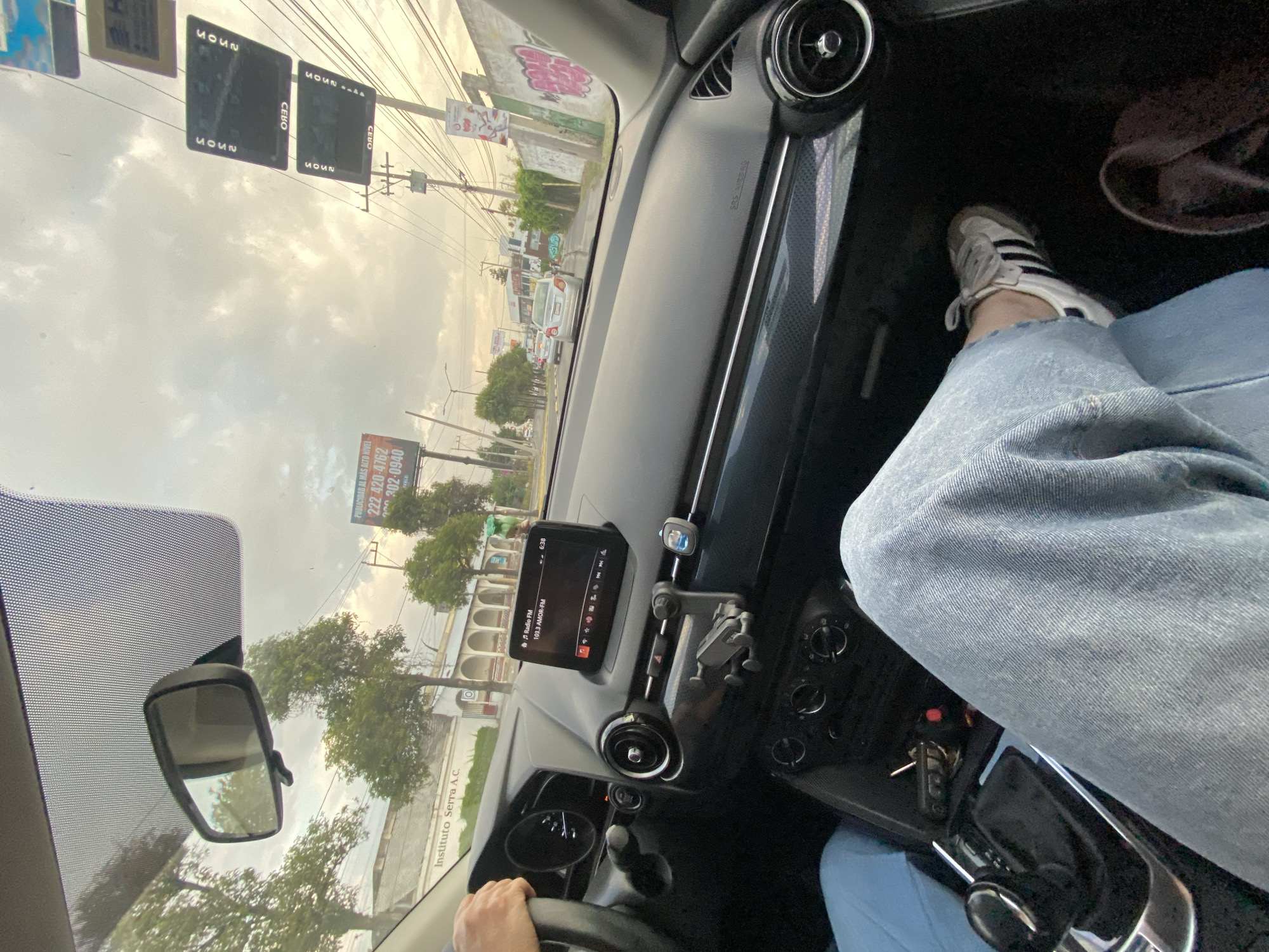 Here is a description of the image:
The image shows a view from inside a car, likely the driver's perspective. The dashboard is visible, including the steering wheel, infotainment screen showing the radio playing, and air vents. A person's leg wearing jeans and a white Adidas sneaker is visible on the right side of the frame. Through the windshield, a street scene is visible with buildings, trees, power lines, and advertising signs. One sign is for "Instituto Serra A.C." and another includes the phone number "222 420-4762". Street signs indicate "CEBO" and "MOUN". It is daylight, with overcast skies.