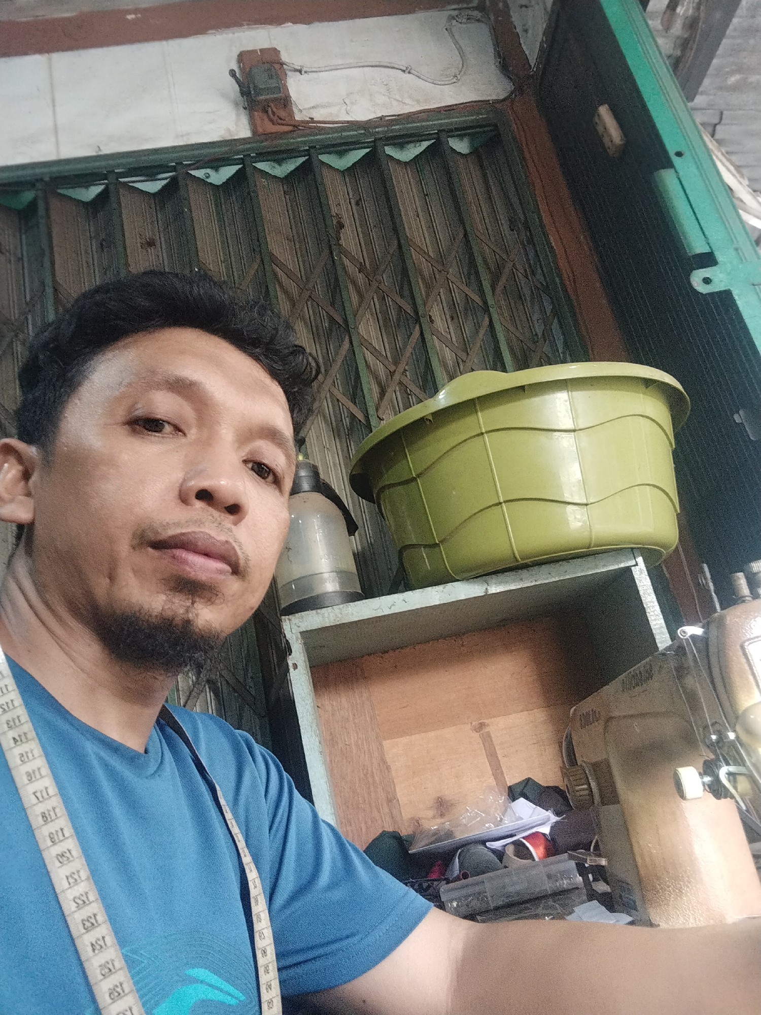 The image is a selfie taken by a man with dark, curly hair and a goatee, wearing a blue t-shirt and a measuring tape draped around his neck. He is in what appears to be a tailor's shop. In the background, there is a collapsible metal door, a green plastic basin on a shelf, a large glass water dispenser, and a sewing machine. The environment suggests a commercial or workshop setting. The overall impression is a candid moment captured by the individual, likely in his place of work. The location is identified as Unknown, Indonesia.