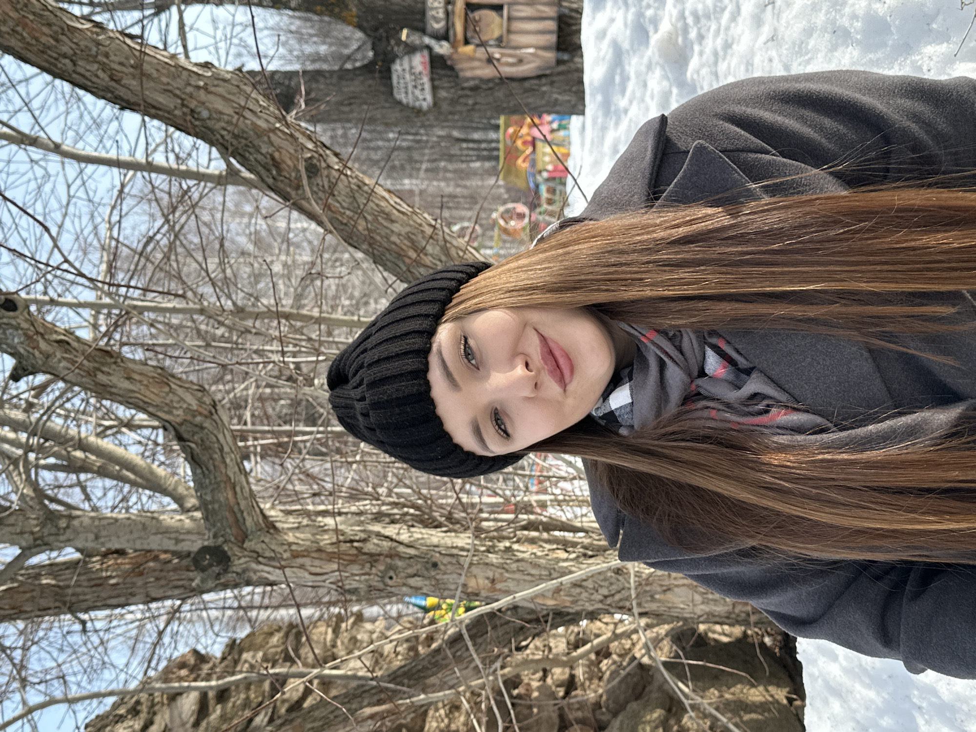 A female subject is captured from the waist up in a snow-covered outdoor setting, identified as the Park of Fairy Tales (Skazka Park) in Chaikovsky, Russia. She is positioned centrally, gazing directly at the camera with a slight smile. Her long, straight, reddish-brown hair cascades over her shoulders and chest. She is dressed in a black ribbed knit beanie, a dark grey wool coat, and a grey and white plaid scarf with thin red lines. The background consists of bare deciduous trees against a clear blue sky. To the left, a large tree trunk with rough bark and a possible rock formation is visible. To the right, a rustic wooden birdhouse or similar structure is attached to a tree trunk, with a light-colored sign above it. In the mid-ground, behind the snow-covered terrain, colorful playground or decorative park elements, including a carousel-like structure, are discernable. The ground is uniformly covered in white snow.