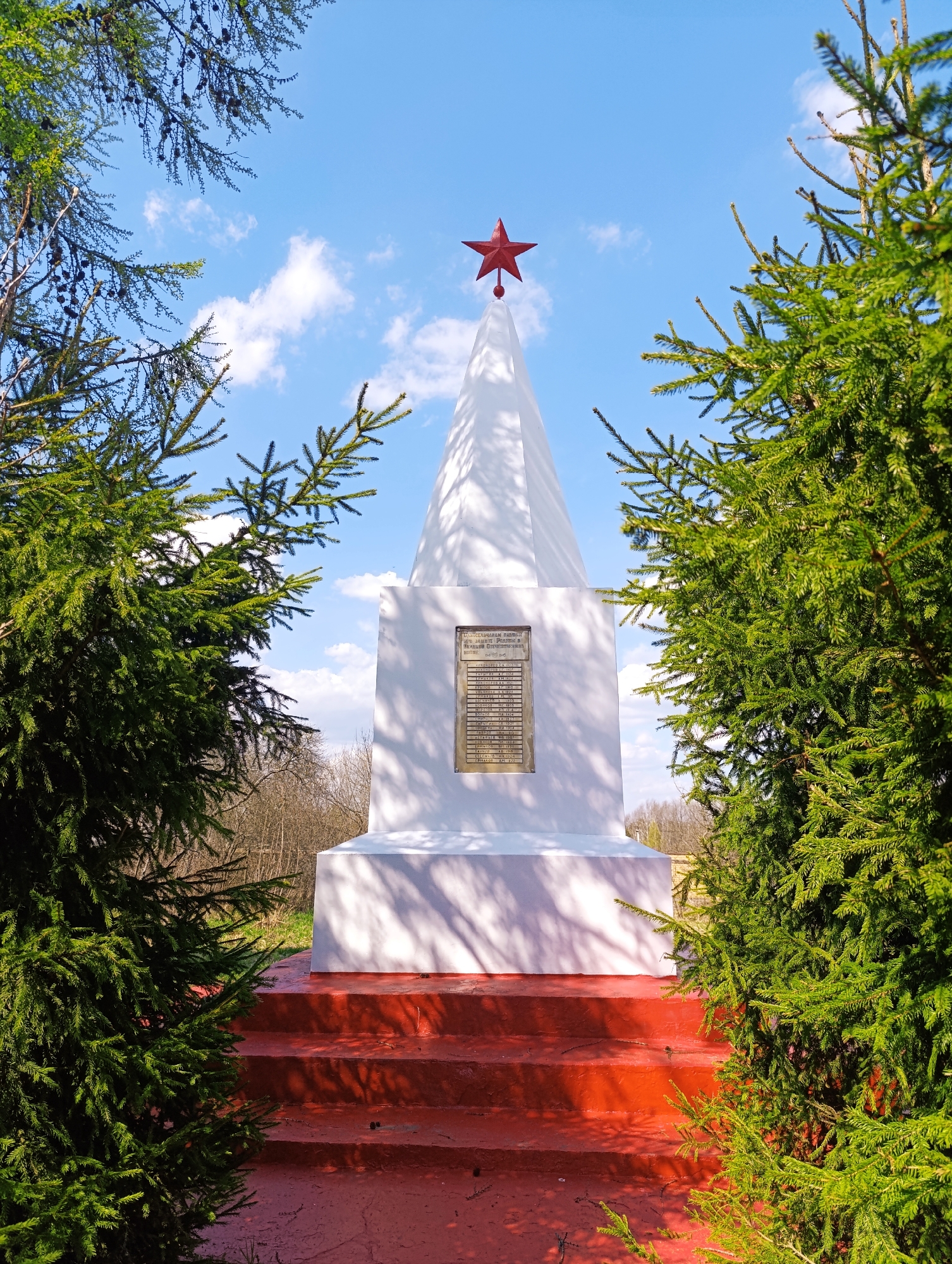 The image shows a white obelisk-style monument with a red star on top, viewed between two evergreen trees. The monument has a plaque on its front. The base of the monument is on red stairs. The background shows a blue sky with scattered white clouds. A small patch of barren trees is visible behind the monument. The location is specified as Ivanovskoe, Russia.