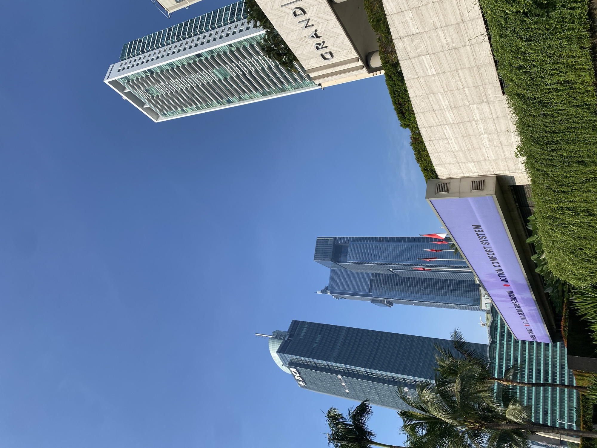 The image shows a view of the skyline in Central Jakarta, Indonesia. Several modern skyscrapers are visible against a clear blue sky, including the BCA Tower. A sign reads "MOTION COMFORT SYSTEM" and "MULTILINK REAR SUSPENSION." Palm trees are in the foreground. A building labeled "GRANDI" is partially visible at the top. Lush greenery is also present.