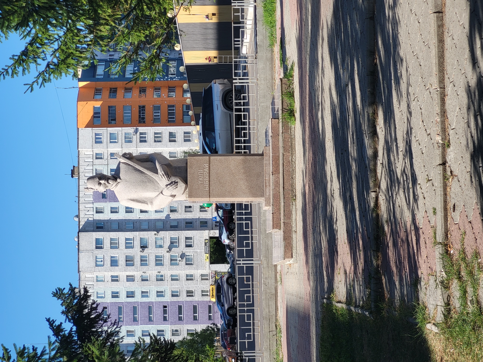 This is a full-shot, eye-level, outdoor, daytime photograph of a stone monument in a city square in Petropavl, Kazakhstan. The monument is dedicated to Mukhtar Auezov and is located in front of a colorful, multi-story apartment building. The monument is made of light-colored stone and features a statue of Auezov holding a stringed instrument. The base of the monument has text inscribed on it. Behind the monument, the apartment building is visible, with different sections painted in shades of grey, purple, orange, and dark blue. The building has many windows. In front of the building, a street with cars is visible, separated from the square by a decorative metal fence. The square itself is paved with interlocking bricks, and there are patches of grass and trees on the left and right sides of the frame. The sky is clear and blue.