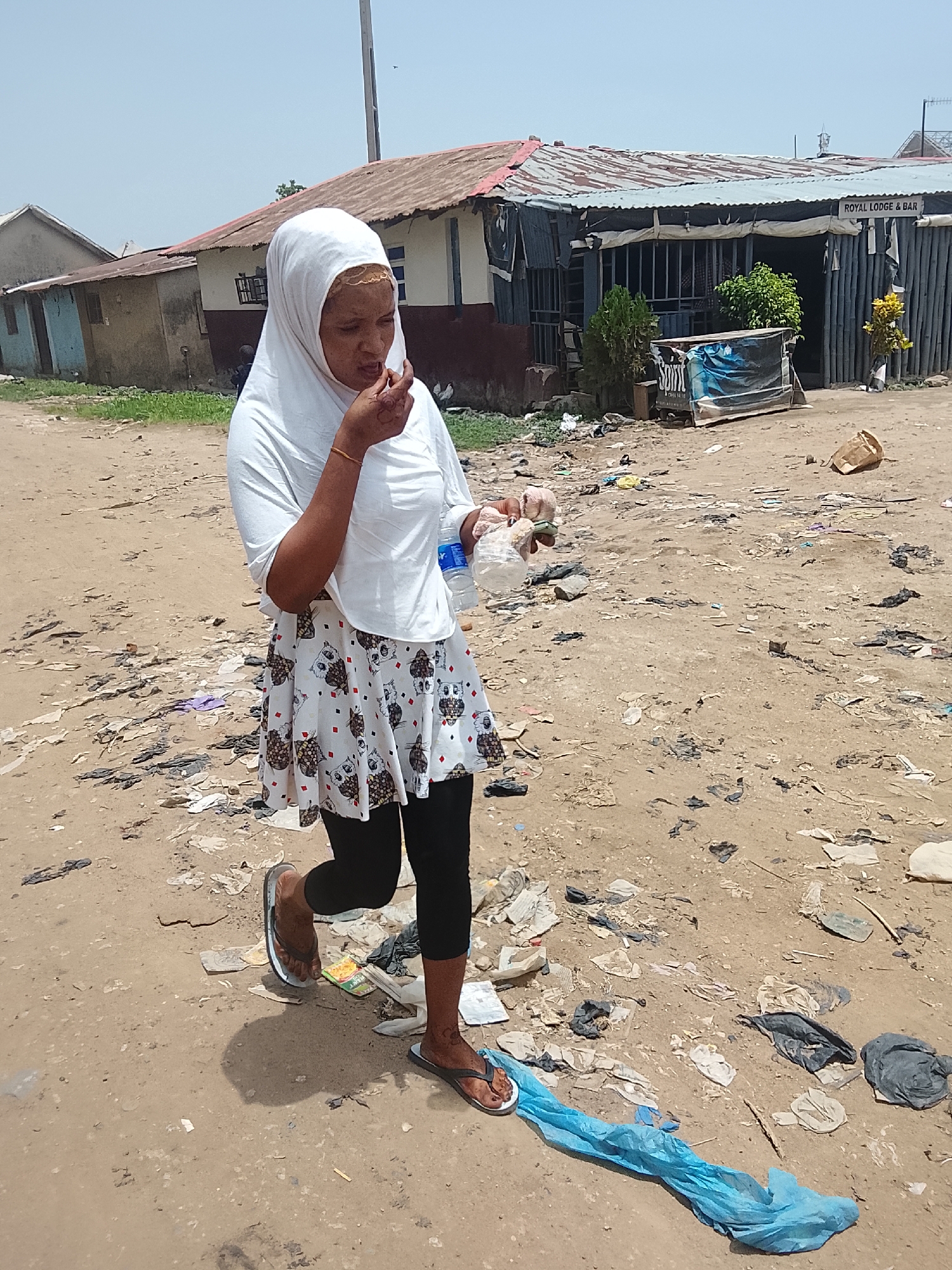 A woman wearing a white hijab, a white owl-print skirt, and black leggings walks on a dirt road littered with trash. She is eating something from a clear plastic bag and holding a water bottle. Buildings are in the background, one labeled "ROYAL LODGE & BAR". The location is in Abuja, Nigeria.