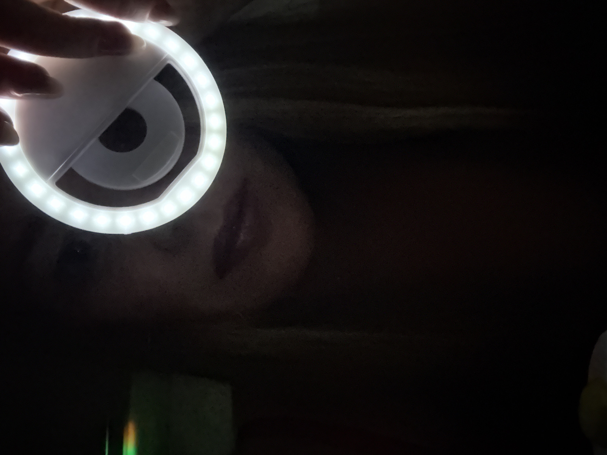 The image captures a close-up, low-light scene featuring a person using a circular LED ring light. A hand is visible at the top, holding the white ring light, which emits bright illumination from its numerous small LEDs arranged in a circle. Through the center of the ring light, a dark, circular lens, likely from a smartphone or camera, is visible.
Part of a person's face, specifically the mouth with what appears to be lipstick and the chin area, is dimly lit by the ring light from below, suggesting the light is clipped onto a device pointed at the subject. strands of hair are also visible in the upper right background. The surrounding environment is predominantly dark, indicating the photo was taken in a dimly lit room or at night, with the ring light serving as the primary source of illumination. The activity depicted is likely the person taking a selfie or recording a video. No text is visible in the image. The location context provided is Chaikovsky, Russia.