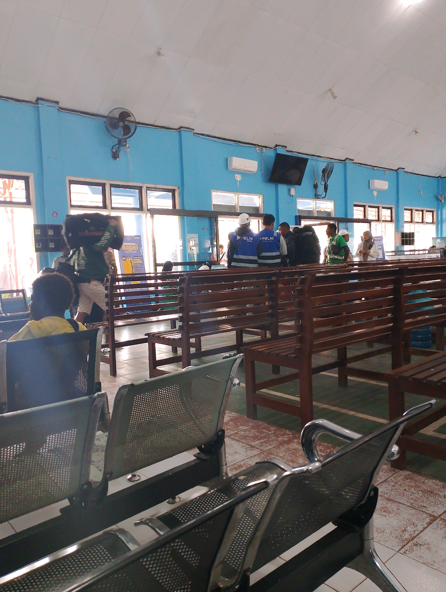 The image depicts the interior of a waiting area, likely at a ferry terminal or transportation hub, in Merauke, Indonesia. The walls are painted a bright blue, and the ceiling is white, with a tiled floor. Several rows of wooden benches fill the space, some with metal chairs interspersed. Natural light streams in through large windows along one side of the room.
In the foreground, several metal chairs with perforated seats are visible. In the middle ground, a person wearing a yellow shirt is seated, facing away from the camera. Behind them, more wooden benches are occupied by people waiting. A man carrying a large dark object on his head walks through the area. Several other individuals, some wearing blue vests with "PELNI" printed on them, are gathered, possibly staff members.
The background shows more windows and a blue wall with what appear to be air conditioning units and a television screen mounted on the wall. A ceiling fan is also visible on the blue wall. The overall impression is one of a busy but orderly waiting area.