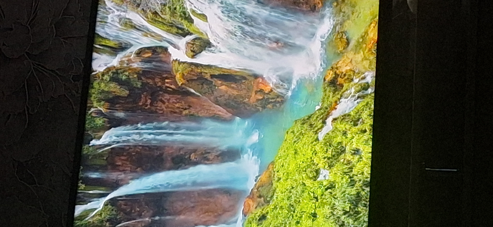 The image shows a scenic view of a multi-tiered waterfall cascading down rocky cliffs covered in lush green vegetation. The water is a vibrant turquoise color, and the surrounding rocks are a mix of brown and orange hues. The foreground features a dense carpet of bright green foliage, likely moss or small plants, with some smaller streams of water trickling down. The overall impression is one of natural beauty and tranquility. There are no people or man-made objects visible in the image.
It is not possible to determine the location in the city of Bila Tserkva, Ukraine from this image, as it depicts a natural landscape without any identifiable landmarks or urban features.