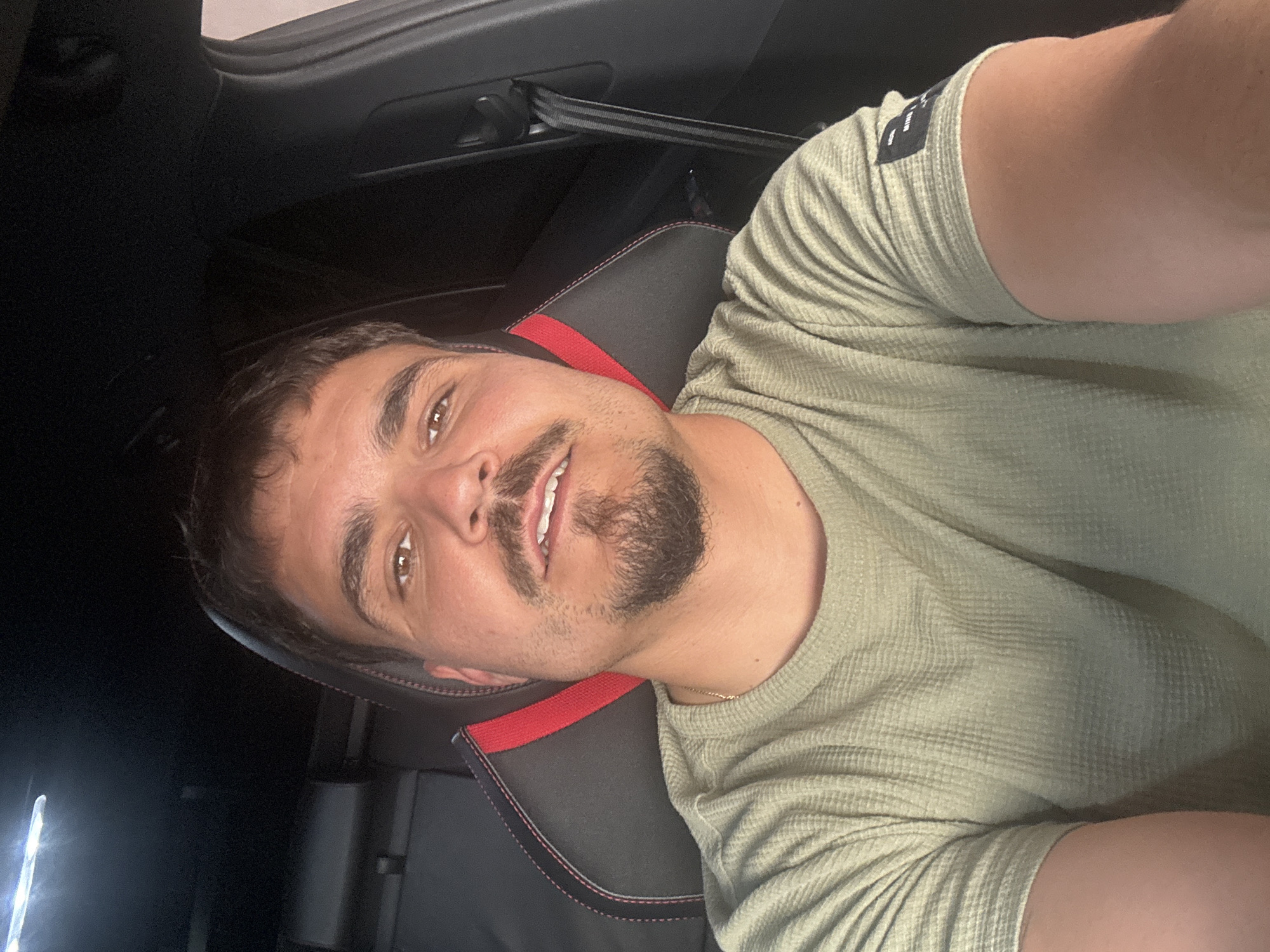 A man with brown hair and a beard is sitting in the driver's seat of a car. He is wearing a light green shirt. The car seat is black with red trim. The car is located in the city of Unknown, Unknown.