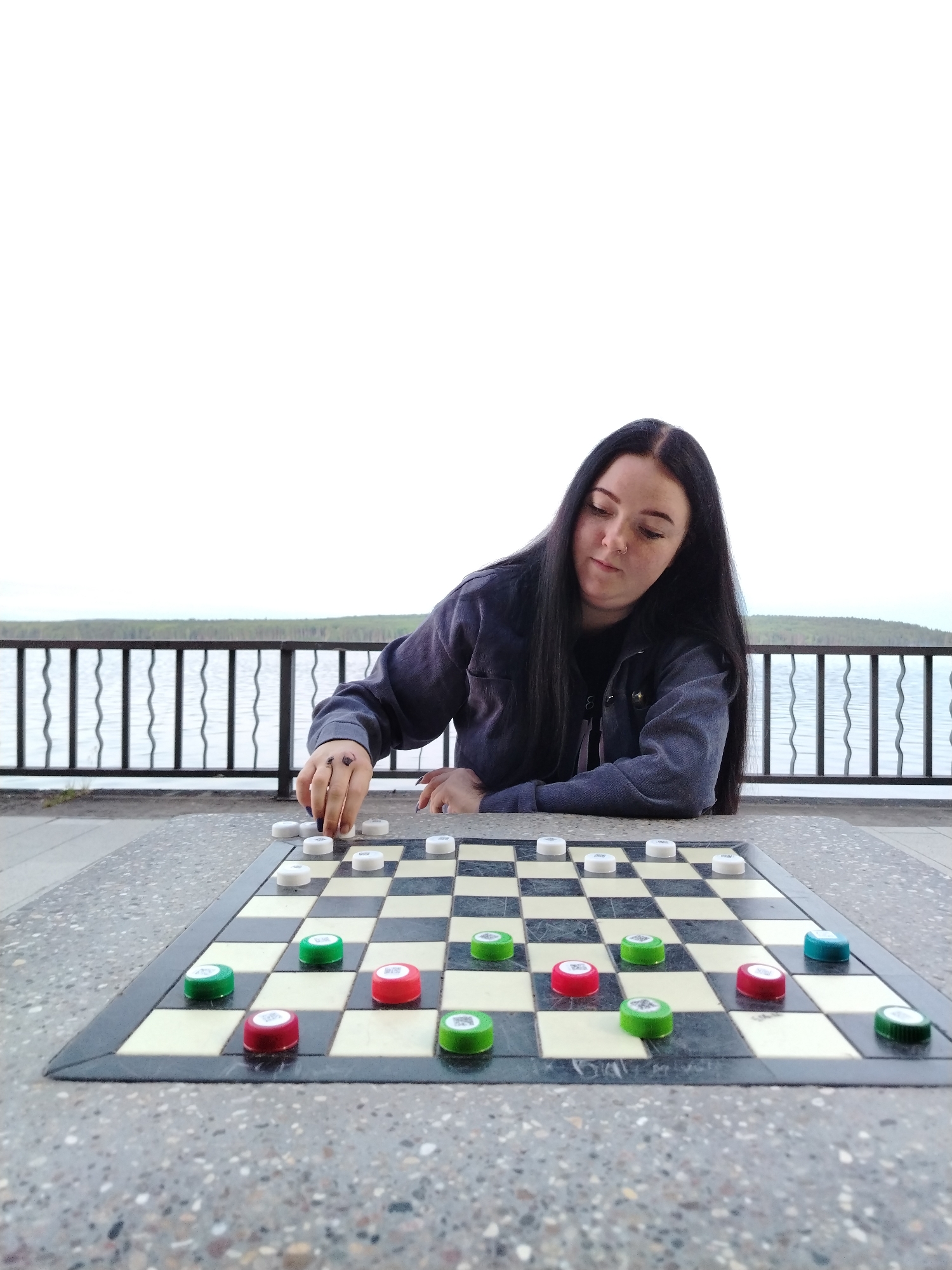 In Chaikovsky, Russia, an outdoor scene depicts a young adult female with long, dark hair, wearing a dark shirt and a blue-grey denim jacket, seated at a speckled grey concrete table. Her left hand manipulates an inverted white bottle cap, serving as a game piece, on an inlaid checkerboard. The opposing game pieces consist of green and red bottle caps, with one teal cap also present. Behind the table, a black metal railing with decorative wavy vertical bars overlooks a expansive body of water, likely the Kama River or Votkinsk Reservoir, under an overcast sky. A distant tree-lined horizon is visible across the water.