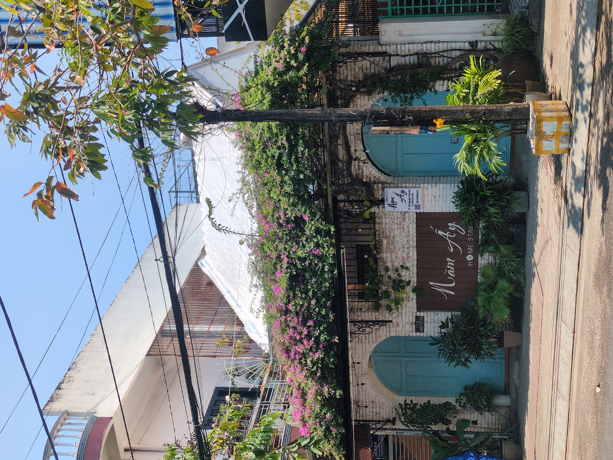 The image depicts the exterior of a building identified by a wooden sign as "Năm Ay Home Stay," located at 118 Nguyễn Thái Học in Tuy Hòa, Vietnam. The building features a white brick facade, with a distinctive arched turquoise-colored door. Bougainvillea plants with vibrant purple flowers cover a significant portion of the building's front and the adjacent pole. A white tarp is visible above, providing shade. The scene is brightly lit, suggesting a sunny day. A tree with green and orange leaves partially obscures the upper left corner of the image. Utility wires crisscross the sky above the building. The sidewalk is paved with reddish-brown tiles, and there are several potted plants placed in front of the entrance. A small sign with the text "NESTAY" is visible next to the front door, along with a QR code. A painted pole wrapped in tape is on the sidewalk.