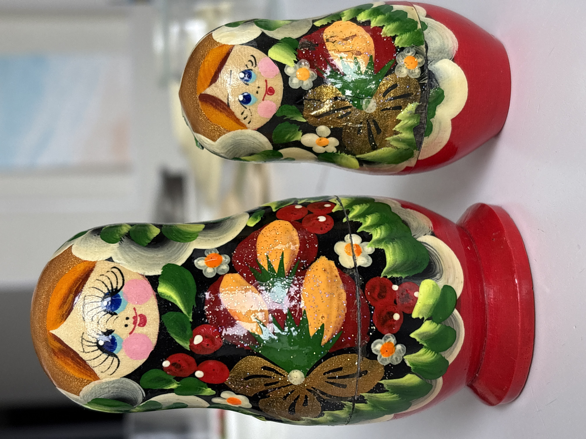 The image displays two vibrantly hand-painted wooden Matryoshka dolls, also known as Russian nesting dolls, lying horizontally on a light-colored surface. The dolls are oriented with their faces pointing towards the left of the frame, showcasing their intricate details.
Each doll is elaborately decorated in a traditional Russian folk art style. Their faces feature large, expressive blue eyes framed by long, dark eyelashes, rosy pink cheeks, and small red lips, conveying a cheerful and innocent expression. Their hair is painted in shades of brown and gold.
The bodies of the dolls are adorned with intricate floral and fruit patterns set against a dark, likely black, background. These designs include lush green leaves, clusters of bright red berries or cherries, and small white flowers with orange centers. Prominent golden-brown decorative elements, possibly representing bows or stylized plants, are embellished with sparkling glitter, adding a shimmering effect to the artwork. Both dolls have a distinctive red base.
The upper doll appears slightly smaller than the lower one, characteristic of a set of nesting dolls. The background is softly blurred, indicating an indoor setting and ensuring the detailed craftsmanship of the dolls remains the focal point. The clear lighting enhances the rich colors and the sparkle of the glitter. These traditional dolls are a strong cultural representation from Russia, aligning with the provided context of Ulyanovsk. There is no visible text in the image.