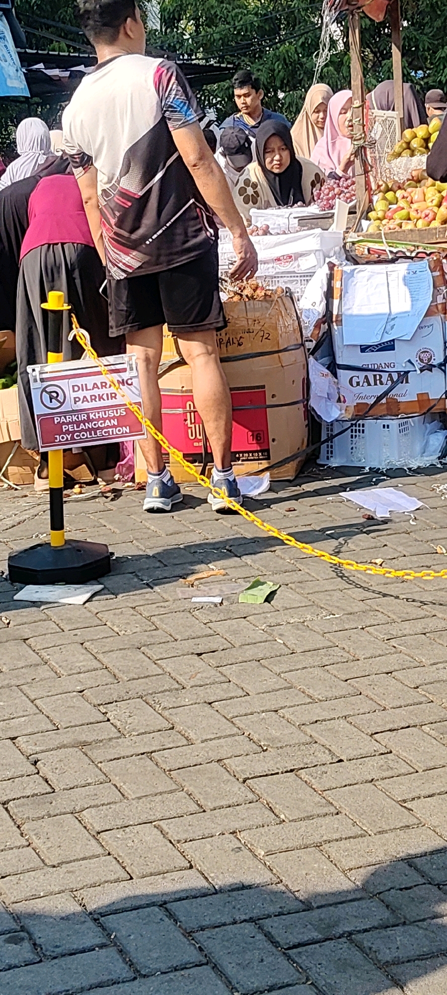 In the city of Cibatu, Indonesia, an outdoor market scene unfolds under a clear sky. The foreground is dominated by a man wearing a black and white t-shirt and black shorts, his legs casting a shadow on the interlocking brick pavement. A yellow chain fence cordons off an area marked with a "DILARANG PARKIR" (No Parking) sign, indicating "PARKIR KHUSUS PELANGGAN JOY COLLECTION" (Parking for Joy Collection Customers Only). Behind the man, several people are visible, including women in hijabs and a young man. Stalls are laden with goods, including piles of fruits like grapes and what appear to be apples or similar produce, alongside stacked boxes and large cylindrical containers. One box is marked with "GARAM International" and another with "J U" and "16". The overall impression is a busy, commercial environment on a sunny day.