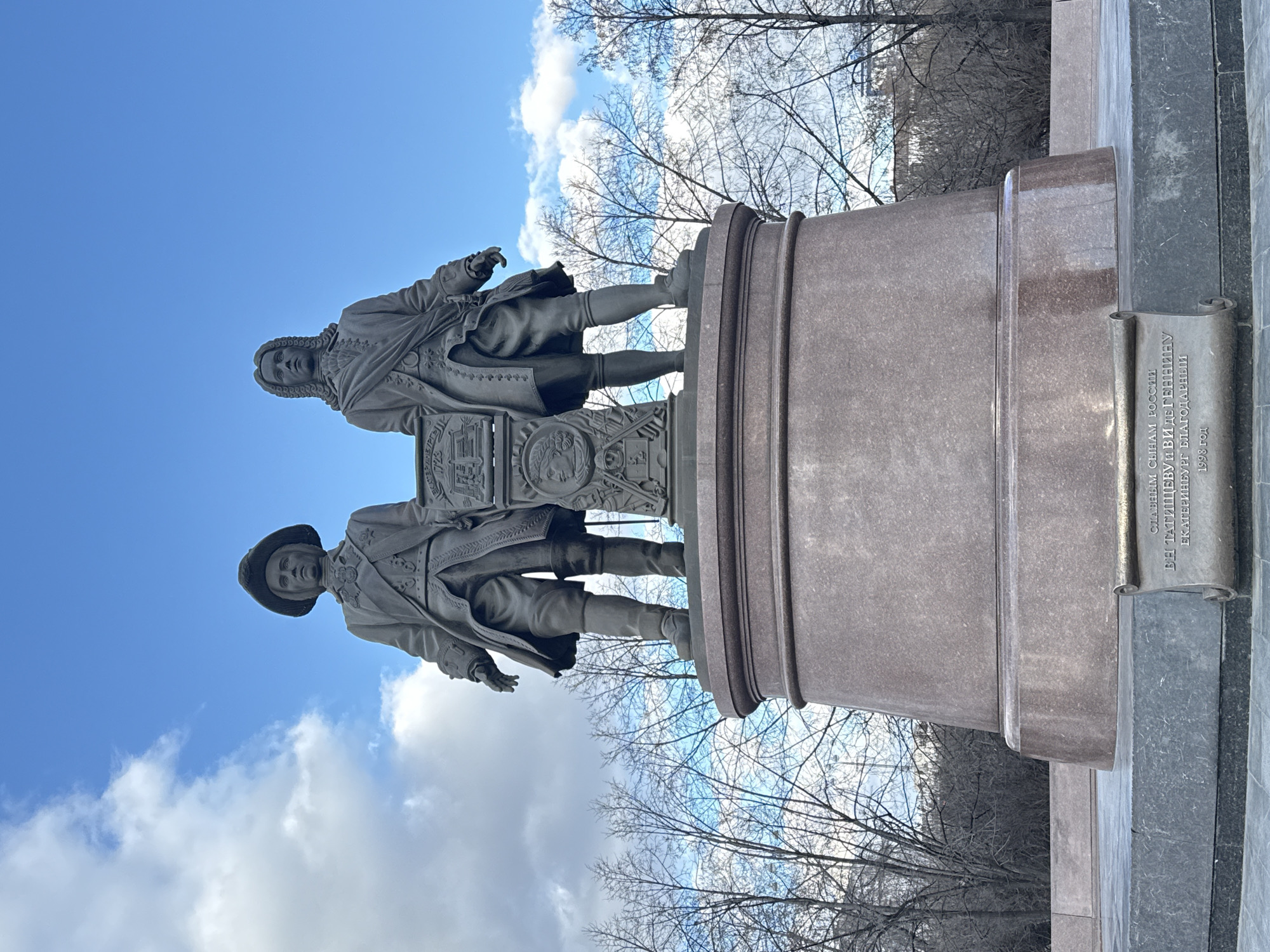 The image depicts a monument in Yekaterinburg, Russia, specifically the monument to Vasily Tatishchev and Wilhelm de Gennin. The monument features two figures in 18th-century attire standing on a pedestal. The pedestal is made of dark stone. A plaque is attached to the side of the pedestal. The monument stands against a backdrop of a blue sky with white clouds and bare trees.