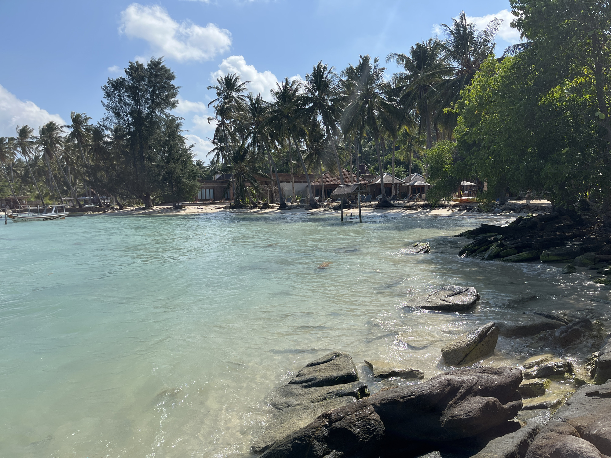 The image depicts a tropical beachfront scene in Legonipah, Indonesia, under a bright, partly cloudy sky. The foreground is dominated by clear, shallow turquoise water lapping against a rocky shore with dark, weathered stones. Beyond the water, a sandy beach stretches into the distance, lined with numerous palm trees. Several small, traditional-style buildings with thatched roofs and open-air seating areas are visible along the beach. In the background, more dense vegetation and palm trees create a lush, green backdrop. A white boat with dark trim is moored near the left side of the frame. The overall impression is one of a serene, tropical resort or secluded beach location.