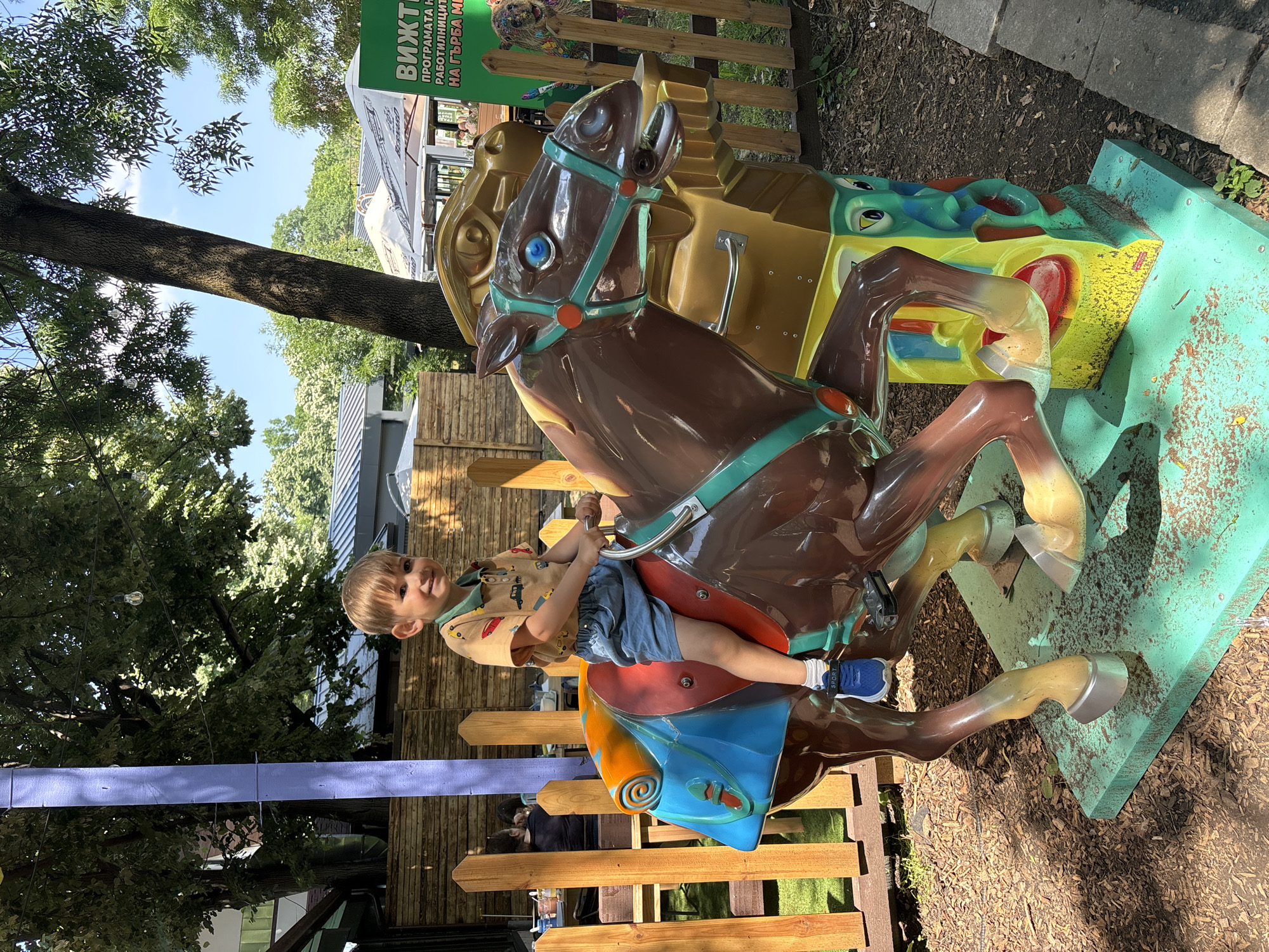 This image depicts a young boy seated on a coin-operated carousel horse in an outdoor recreational area within the city of Sofia, Bulgaria.
In the center, a fair-skinned young boy with light brown hair, appearing to be a toddler, sits on a brown plastic carousel horse. He is wearing a light brown collared short-sleeved shirt patterned with small colorful cars, light blue shorts, white socks, and blue sneakers with "SPORT" written on them. The boy is smiling directly at the camera while holding onto the silver handlebars of the horse ride.
The carousel horse is predominantly dark brown with teal-colored reins and bridle. Its eyes are blue. The saddle is sculpted in shades of blue, orange, and red. The horse stands on a teal-green rectangular base, which shows signs of wear and discoloration. Beneath the horse, part of the mechanical structure is visible, colored in yellow and turquoise with painted cartoonish eyes and features.
The immediate ground around the carousel is covered with dark brown wood chips or mulch. To the left, a low wooden fence separates the boy and the ride from an outdoor seating area. Behind the fence, a person with dark hair, wearing a dark shirt, is seated at a wooden table facing away from the camera. A tall, light purple vertical post is prominent behind the fence on the left side.
To the right of the carousel horse, the wooden fence continues. Behind this section of the fence, a large green vertical sign with Cyrillic text is partially visible. The text clearly states "ВИЖТЕ ПРОГРАМАТА НА РАБОТИЛНИЦИТЕ НА ГЪРБА МИ," which translates to "SEE THE PROGRAM OF THE WORKSHOPS ON MY BACK," indicating details for activities. To the right of the sign, a colorful, abstract animal sculpture, possibly a squirrel or marmot, is perched behind the fence.
The background features dense green foliage from large trees, providing substantial shade. String lights are visible hanging between some trees. Several white canopies or large umbrellas cover outdoor seating areas, and parts of buildings with dark roofs and windows are discernible further back. The sky above is clear blue with minimal cloud cover. Paving stones are visible on the far right edge of the ground.