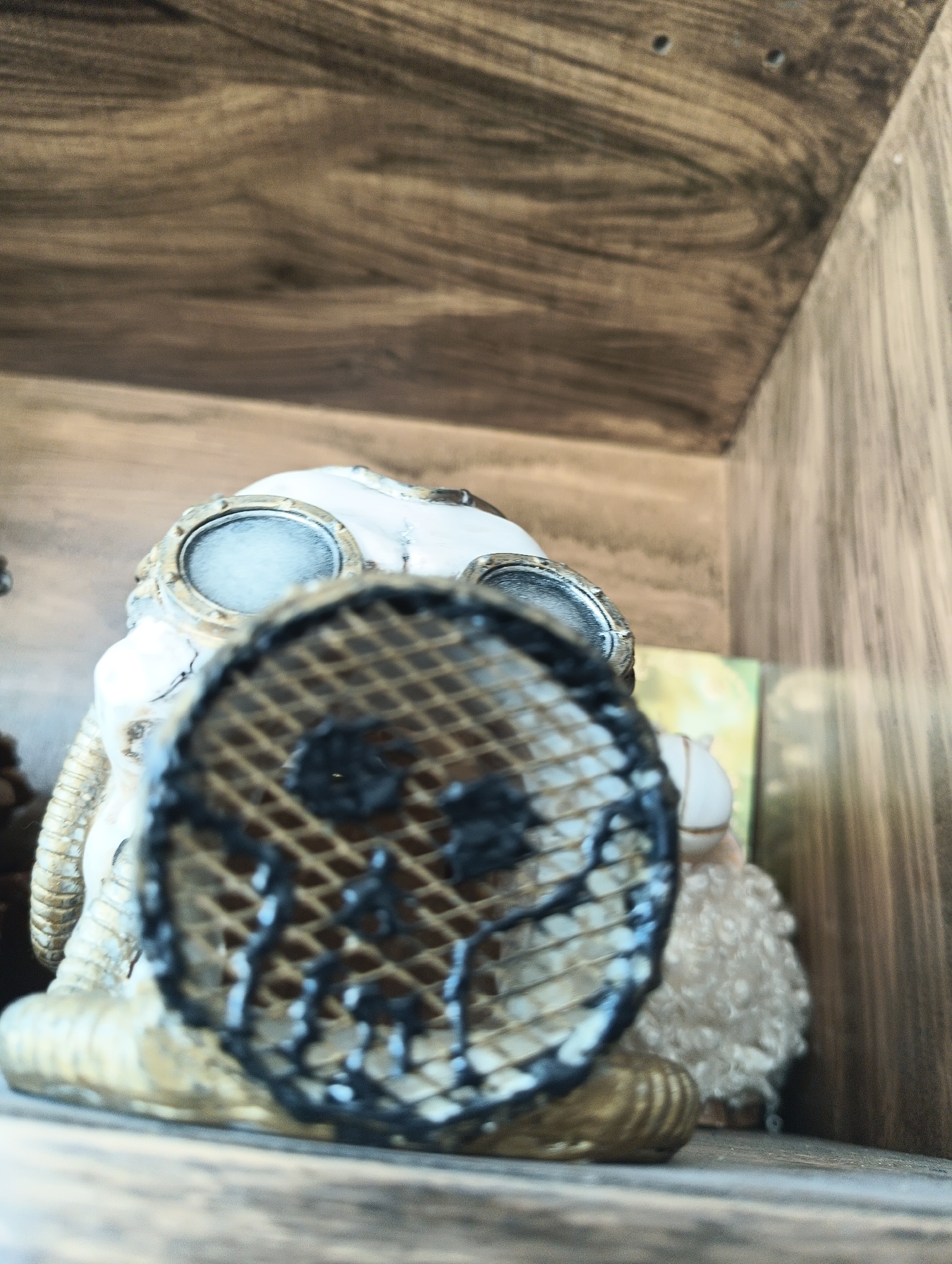 The image shows a close-up, low-angle shot of a doll or figurine with a gas mask. The figurine has a white, skull-like head with large, round, metallic goggles. A dark, grid-like mask covers the front of the face, obscuring the features underneath. The figurine appears to be made of resin or a similar material, with metallic accents on the goggles and a ribbed tube extending from the side of the head. The figurine has a fluffy, light-colored body.
The figurine is placed on a wooden shelf or surface. The background is out of focus and shows the texture of wood grain, suggesting a shelf or cabinet interior. There is a hint of a colorful object with green and yellow tones behind the figurine. The lighting is soft, and the overall mood is somewhat eerie or mysterious due to the gas mask. No specific activity is depicted; the figurine is stationary. There is no visible text in the image. The location context provided is Karnobat, Bulgaria, but there are no visual cues within the image to confirm this location. The time of day and weather are not discernible.