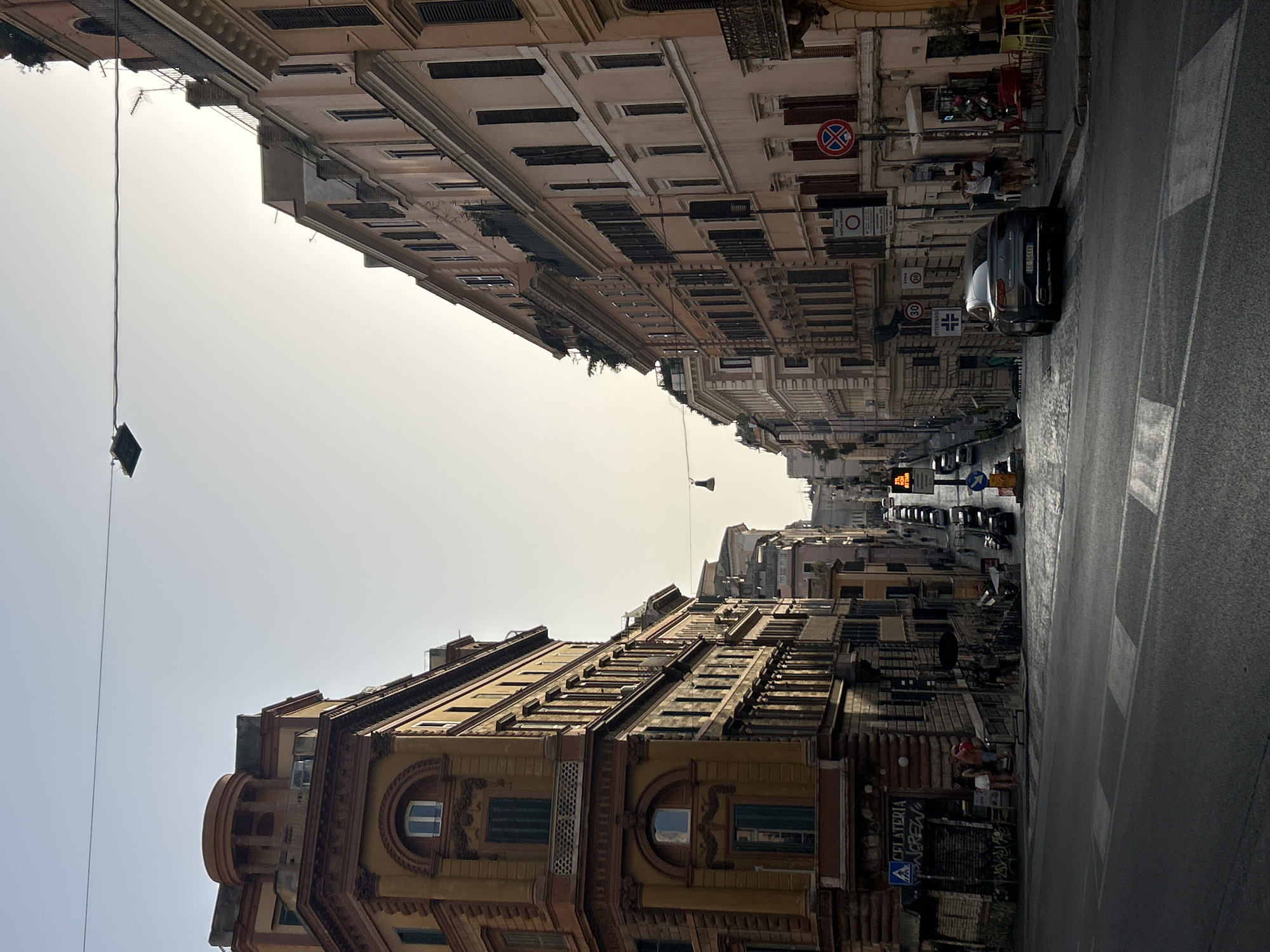 This is a street-level, medium-shot photograph taken in Rome, Italy, looking down a sloping street lined with tall, multi-story buildings on both sides. The sky is a pale, hazy yellow, suggesting either early morning or late afternoon light, with a thin horizontal wire visible across the top of the frame.
On the left, a prominent building with an ochre facade features intricate architectural details, including decorative cornices and arched window surrounds. The building's top has a rounded section with columns and is topped with a darker, flat roof. A sign for "GELATERIA" is visible on this side, with a few people gathered nearby on the sidewalk.
The right side of the street is dominated by a pale pink building with numerous windows, many of which are covered by dark shutters. This building also exhibits architectural ornamentation, including balconies with decorative railings. Several signs are mounted on poles along the sidewalk, including traffic signs and a circular red sign with a blue diagonal bar.
A gray car is parked on the right side of the street, facing uphill. Several other vehicles are visible further down the street, also parked and in motion, indicating traffic flow. Pedestrians are present on both sidewalks, moving or standing. The street itself is paved with cobblestones in some areas and asphalt in the foreground, with visible crosswalk markings. A traffic light displaying red is visible in the distance. The overall impression is an urban street scene with the characteristic architecture of Rome.