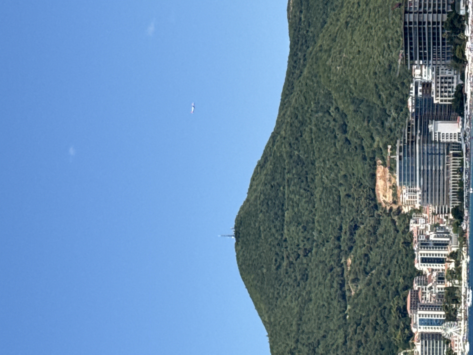 The image depicts a vast expanse of clear blue sky above a densely forested green mountain. A faint, almost imperceptible, distant airplane is visible in the upper right quadrant of the sky. The mountain's peak is topped with a communication tower. At the base of the mountain, a coastal city, identified as Budva, Montenegro, stretches along the shoreline. The city is characterized by a dense concentration of modern buildings, including hotels and residential structures. Some buildings appear to be under construction, with cranes visible on some sites. The foreground shows a body of water with several boats docked, suggesting a harbor or marina. The scene is bathed in bright daylight, indicating a clear and sunny day.