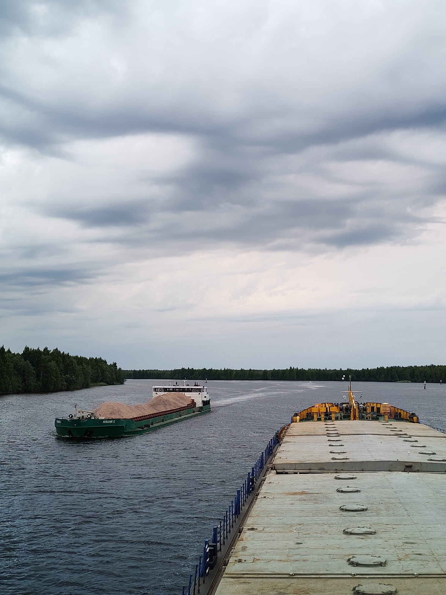 A wide, tranquil river flows under a cloudy, overcast sky, hinting at an approaching storm. The water is a deep, choppy blue, reflecting the muted tones of the sky. On the left bank of the river, a dense forest of green trees stretches as far as the eye can see, creating a natural, verdant border.
Two barges are prominent on the water. The barge closer to the left bank, a long, green vessel, is laden with a substantial load of light-colored gravel or sand, its cargo forming a rounded mound. The name "НЕВСКИЙ G" is visible on its side. This barge is moving away from the viewer, heading towards the distant treeline.
In the foreground, partially obscuring the view of the river and the other barge, is the deck of a much larger vessel. This barge is a pale, weathered gray, with numerous circular hatches visible across its surface, suggesting it is a cargo carrier designed for bulk goods. Its structure is robust and utilitarian, with a blue railing along the visible edge. The deck is clean but shows signs of wear and tear.
The overall scene is one of industrial transport set against a natural landscape. The weather is subdued, with the heavy clouds creating a dramatic and somewhat somber atmosphere. The time of day is likely afternoon or early evening, given the soft, diffused light. The presence of the barges and the cargo indicates ongoing commercial activity on the waterway, likely in the region of Novokemskii, Russia, given the location context. There are no people visible in the frame.