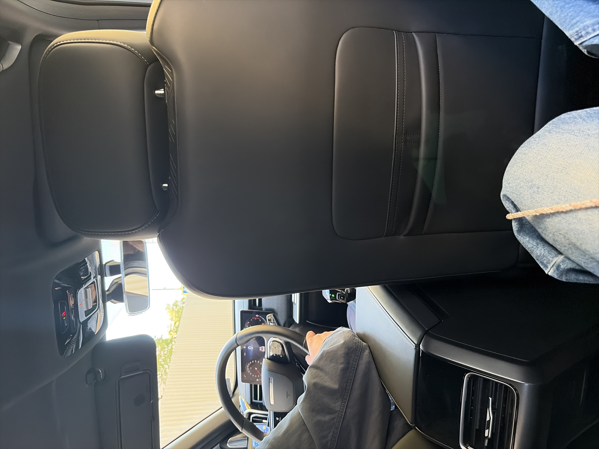The image shows the interior of a car, likely a modern SUV, from a rear passenger's perspective. In the foreground, a person's legs, clad in blue jeans, are visible. The car's interior features black leather seats, a center console with an air vent, and a digital dashboard with the steering wheel in view. The driver's hand is on the steering wheel. The background shows a view through the car windows of a light-colored fence and some trees, suggesting an outdoor environment, possibly a street or parking area. The car's overhead console shows illuminated buttons, with red and orange lights indicating their status. The image provides no specific clues or markers to pinpoint its location in Moscow, Russia, or any other city.