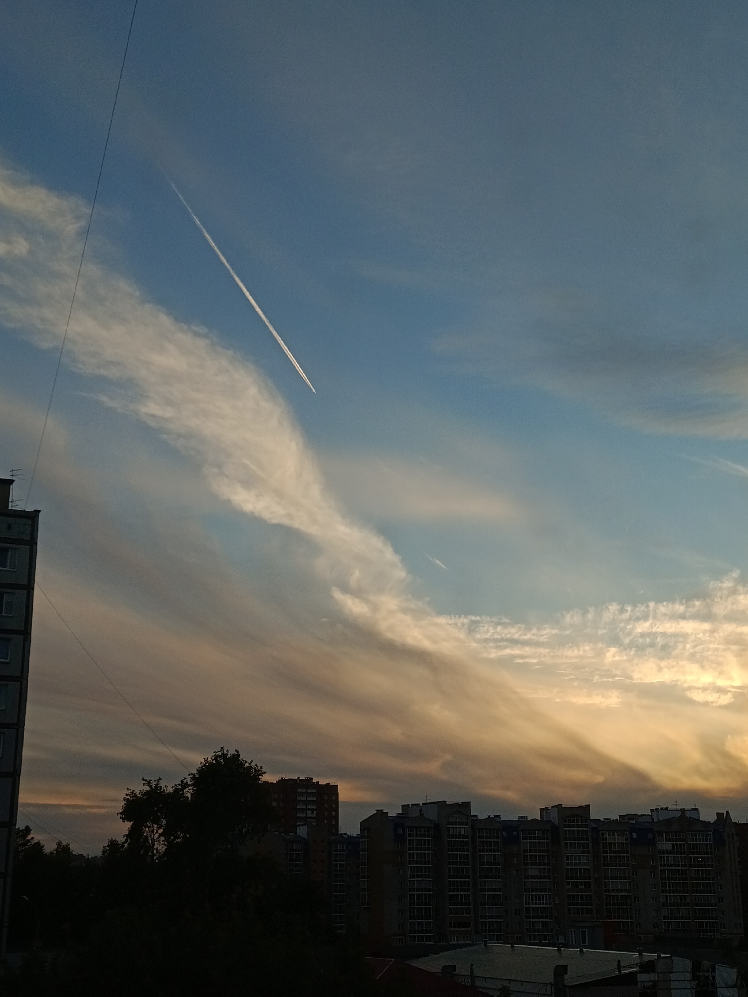 The image depicts a city skyline at dusk, likely in Krasnoyarsk, Russia. A clear sky dominates the upper portion of the frame, with a white contrail from an airplane streaking diagonally from the upper left towards the center. Wispy clouds, tinged with orange and yellow hues from the setting sun, stretch across the sky. In the foreground, a silhouette of a multi-story apartment building with visible windows is on the left side. A dense cluster of trees, also in silhouette, occupies the lower-middle section of the image. To the right of the trees, several more modern apartment buildings are visible, their facades outlined against the fading light. The overall scene captures a quiet urban evening with a prominent airplane trail as a focal point in the sky.