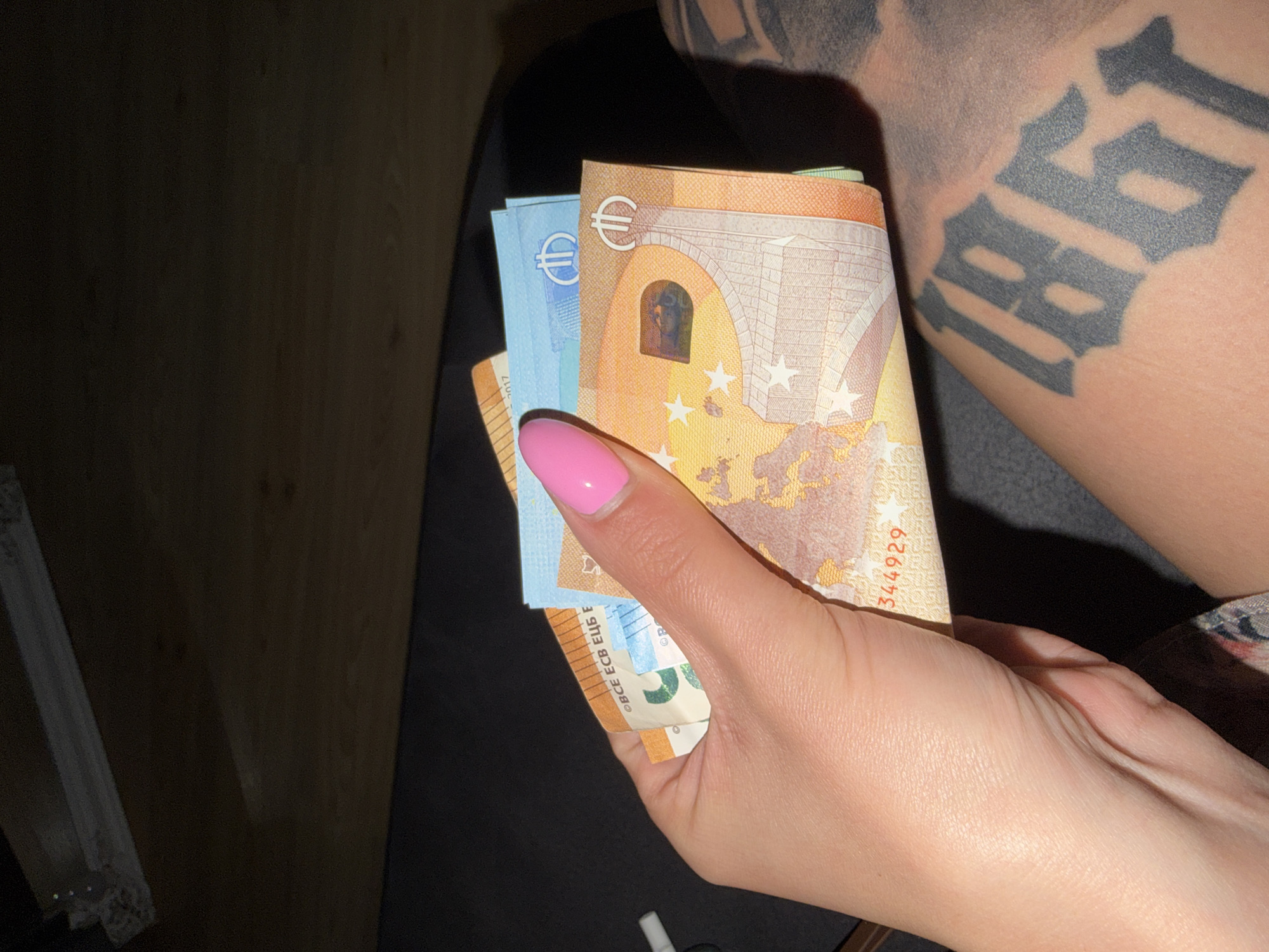 The image captures a close-up of a person's hand holding a fan of Euro banknotes. The prominent note is a 50 Euro bill, displaying its architectural motif and a holographic window. Behind it, a 20 Euro note (blue) is partially visible, and other denominations may be present further back. The hand has light skin and bright pink polished fingernails.
To the right, partially obscured by the hand and notes, is a person's upper arm or shoulder with a dark, block-style tattoo that clearly shows the numbers "187". The background is dark and indistinct, suggesting an indoor setting with subdued lighting.
Visible text on the 50 Euro note includes the Euro symbol (€), "BCE ECB EЦБ E" (indicating the European Central Bank in various languages), and a partial serial number "344929". The 20 Euro note also shows the Euro symbol and "BCE ECB EЦБ E". No specific location cues for Plovdiv, Bulgaria are visible in the image, though the Euro currency implies a European context. The activity depicted is simply holding money. The time of day and emotions are not discernible.