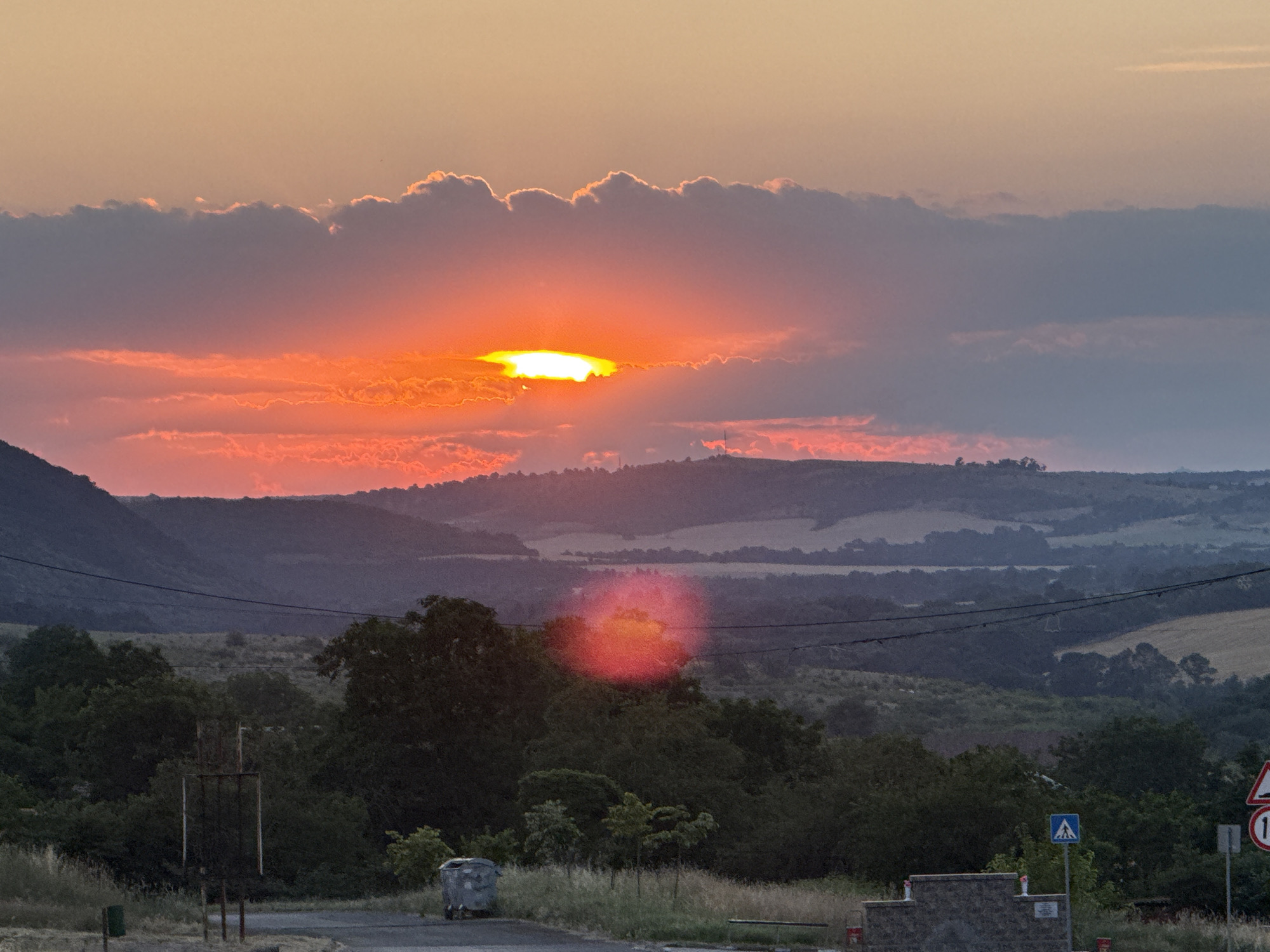 The image depicts a rural landscape in Medovo, Bulgaria, during a sunset. The sky is filled with dramatic clouds, illuminated by the setting sun which is peeking through them, casting a vibrant orange and red glow across the horizon. The landscape features rolling hills covered in vegetation and trees, with a few patches of cultivated fields visible in the distance. In the foreground, a road runs through the scene, with a few objects scattered along its side, including a trash bin, a metal structure, and some road signs. The overall mood of the image is serene and picturesque, capturing the beauty of nature at dusk. The time of day is clearly sunset due to the lighting and colors in the sky. The weather appears clear, with the exception of the clouds which are adding to the dramatic visual. The presence of road signs suggests a populated area, likely a rural road. No people are visible in the frame. The road signs contain symbols and numbers, but no legible text is discernible.