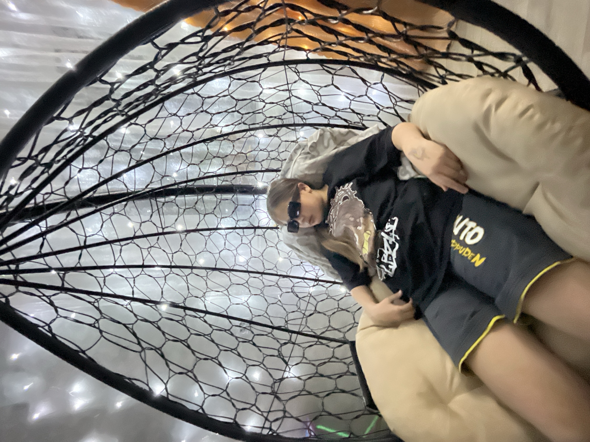 A young person with long hair and sunglasses reclines in a hanging chair, seemingly indoors. The chair's frame is black and woven, adorned with small white lights. The person wears a black long-sleeved shirt, gray shorts with yellow trim, and a light-colored pillow supports them. The background is somewhat blurred. There is no indication of Novopokrovskaya, Russia, in the image.