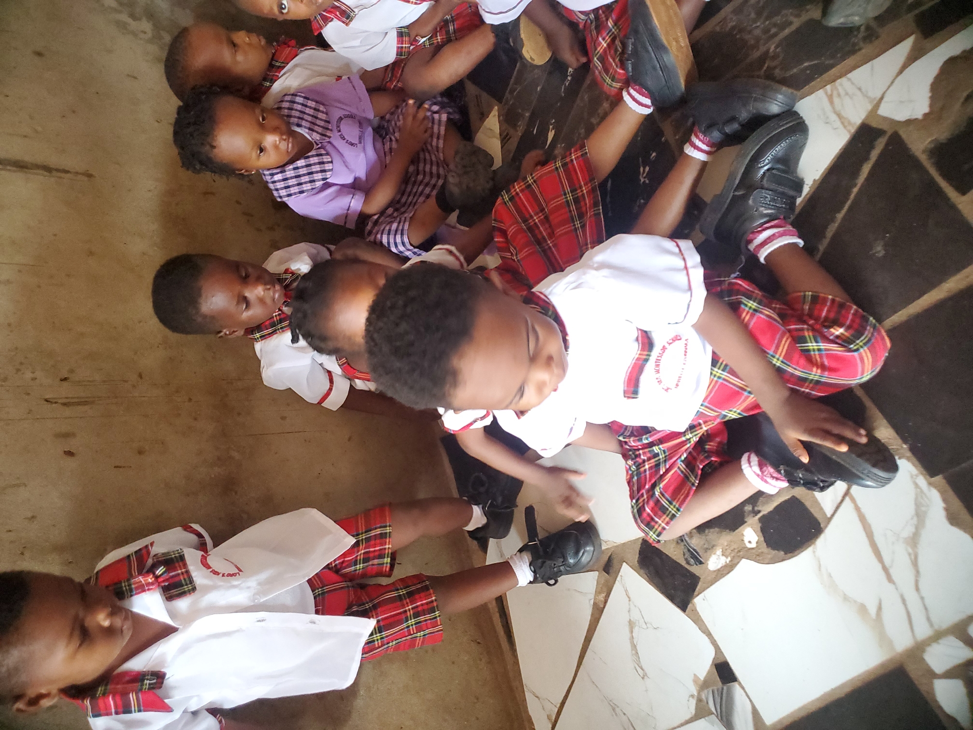 The image captures a group of children in what appears to be an indoor setting in Ukwu-Abwa, Nigeria. The children are dressed in school uniforms, with some wearing red plaid skirts or shorts with white shirts featuring a school logo. One child is wearing a purple dress, also with a logo. The children are sitting or lying on a tiled floor. The lighting suggests an indoor environment, possibly a classroom or common area within a school building. There's minimal background detail visible beyond the floor and the children themselves.