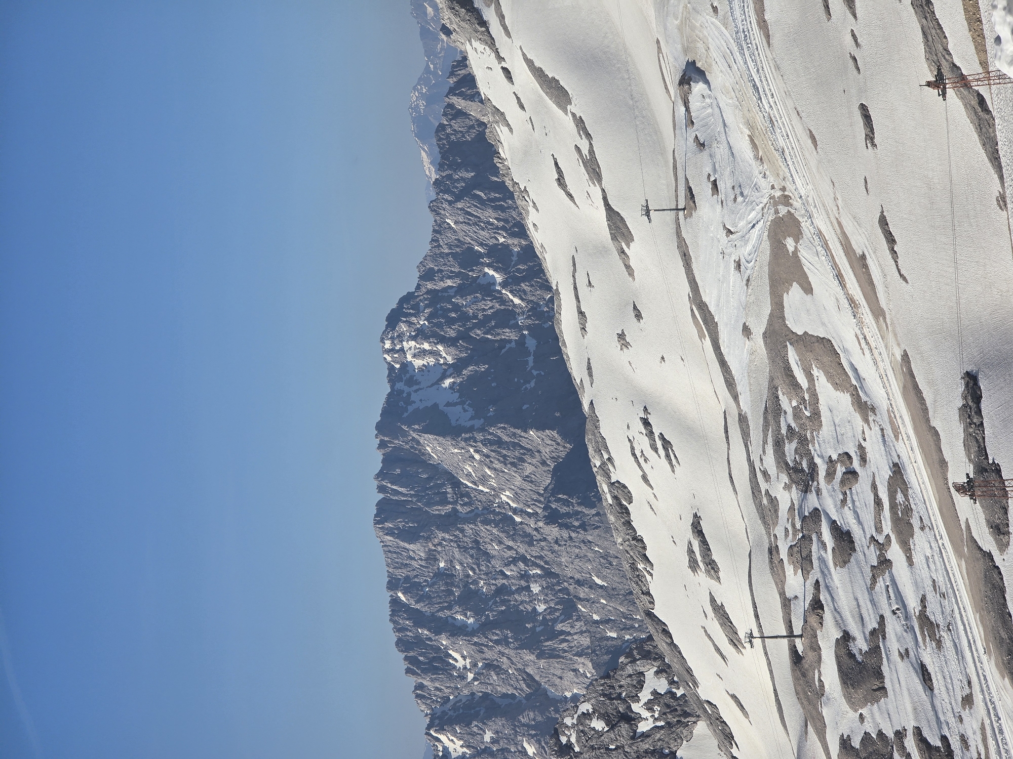 The image shows a vast, snow-covered mountain landscape under a clear blue sky. In the foreground, the snow is patchy, revealing rocky outcrops and ski tracks. Two ski lift towers are visible, one tall and slender, and another shorter, red metal structure. The middle ground features a broad, sloping snowfield with more distinct ski tracks and several more ski lift towers. In the background, rugged, grey mountains rise, their peaks jagged and partially covered in snow. The overall scene suggests a high-altitude location, likely a ski resort, during a sunny day. While the image doesn't explicitly show Garmisch-Partenkirchen, the terrain and presence of ski lifts are consistent with the Bavarian Alps, where Garmisch-Partenkirchen is situated. There are no people visible in the image.