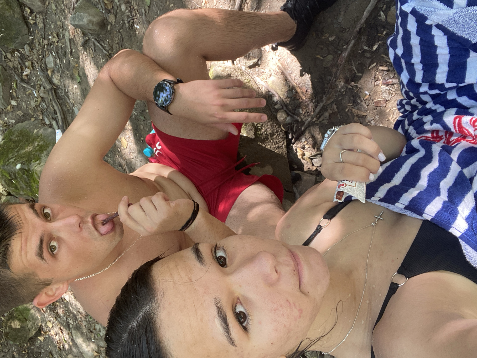 Two individuals, a male and a female, are seated outdoors near a body of water. The male, positioned in the background, is shirtless and wearing red swim trunks. He is holding a stick-like object with his tongue extended towards it. A silver chain is visible around his neck. The female, in the foreground, is wearing a black bikini top and is draped in a blue and white striped towel. She has a silver necklace with a cross pendant. Her right hand, adorned with rings, rests on her lap. The ground is covered with dirt, rocks, and debris. The environment suggests a natural, possibly wooded, setting near Debnevo, Bulgaria.