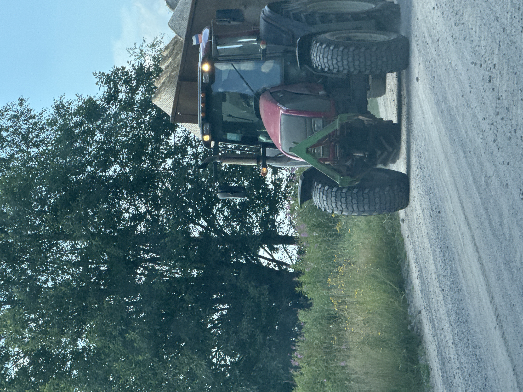 A red tractor with a trailer full of gravel is driving on a gravel road. The tractor has large, rugged tires and is equipped with a front loader. A lush green tree line is visible in the background, with a clear blue sky above. The scene is set in the rural landscape of Yablonovka, Russia.