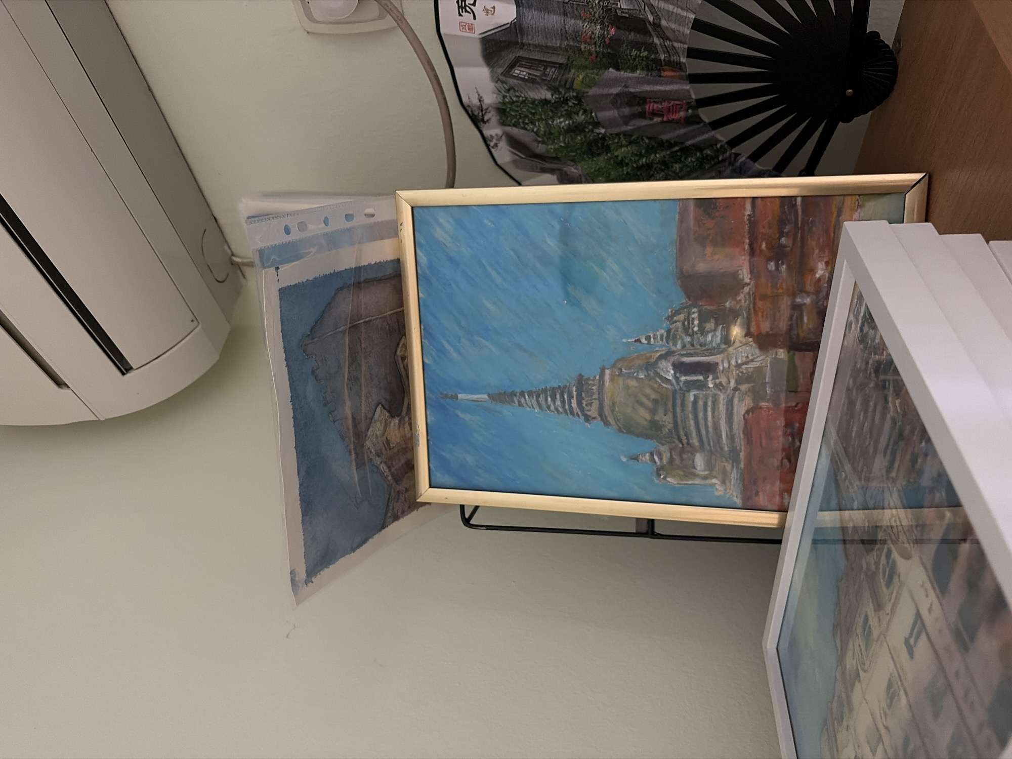 The image depicts a collection of framed artwork and decorative items displayed on a wooden shelf. In the foreground, a white framed artwork is partially visible, showing a reflection of buildings. Behind it, a framed painting of a temple or pagoda with a bright blue sky dominates the scene. To the right of the painting, a black, fanned decorative item with a print of a building and trees is displayed. Above the artwork, a portion of a white air conditioning unit is visible on a light-colored wall. There are no people or discernible actions in the image. The scene appears to be an indoor display of art. The location in Budva, Montenegro is not ascertainable from the image.