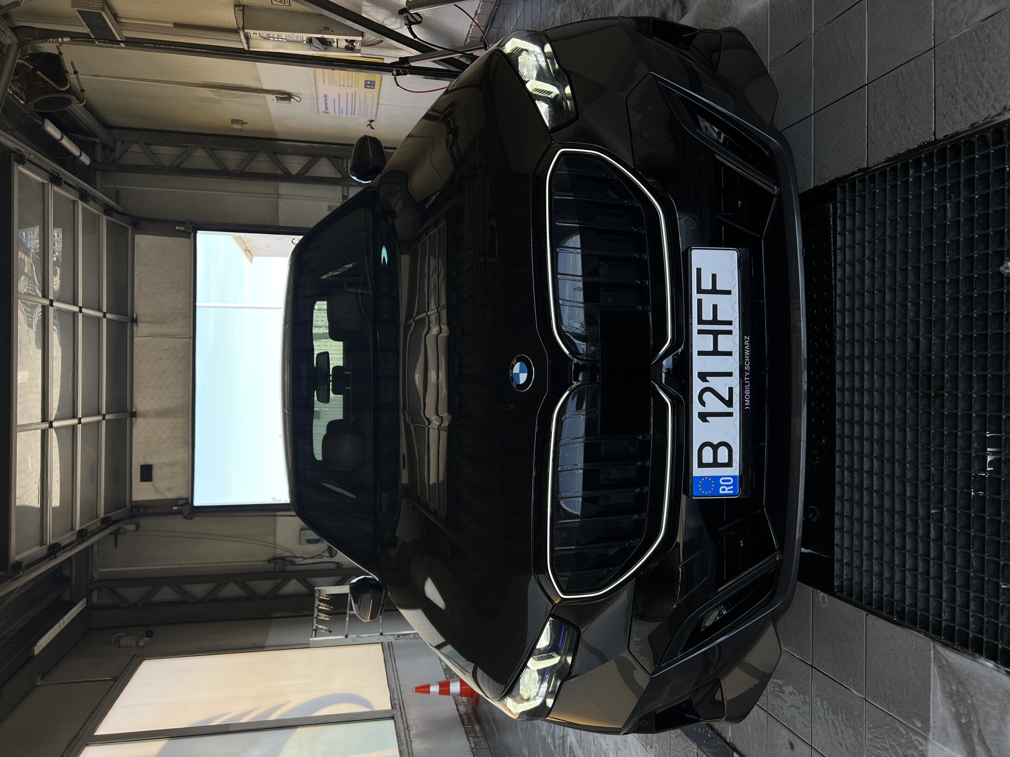 A black BMW is parked in what appears to be a car wash bay. The car is oriented vertically in the frame. The front of the car, including its distinctive grille and headlights, is prominently displayed. A license plate with the characters "B 121 HFF" is visible. Below the license plate, text reads "MOBILITY.SCHWARZ". The car is positioned on a black grated surface, which is common in car wash areas. The background shows a large garage door and a brightly lit opening, suggesting it is daytime. The overall scene is indoors, likely at a commercial car wash facility. There are no people visible in the image. The car looks clean and well-maintained. The lighting suggests it's daytime.
