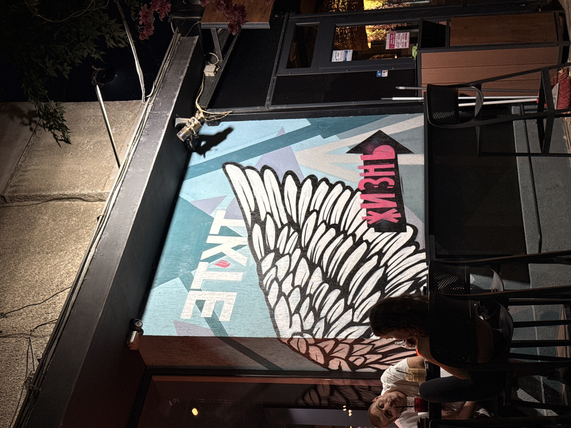 In the city of Lovech, Bulgaria, a night scene unfolds at an outdoor seating area, likely a cafe or restaurant, beneath an overhang. The focal point is a large mural on a wall, featuring a prominent stylized angel wing and geometric patterns in shades of blue and grey. White lettering, "ЭТЛЕ" (ETLE), is visible at the top, and the word "жизнь" (life) is written in pink with an arrow pointing to the right, below the wing.
Two individuals are seated at a table, illuminated by ambient light. A man, wearing a white t-shirt, is on the left, looking towards the camera. A woman, with curly hair, is seated on the right, facing away from the camera. Drinks are present on the table between them.
Above the mural, a black overhang runs horizontally, with various wires and electrical fixtures attached, including a security camera visible on the left. To the right of the mural, a dark-colored doorway leads into a building. Further to the right, a brown trash receptacle and brooms are situated next to the entrance. The environment suggests an urban setting at dusk or nighttime, with the mural serving as a prominent decorative element.