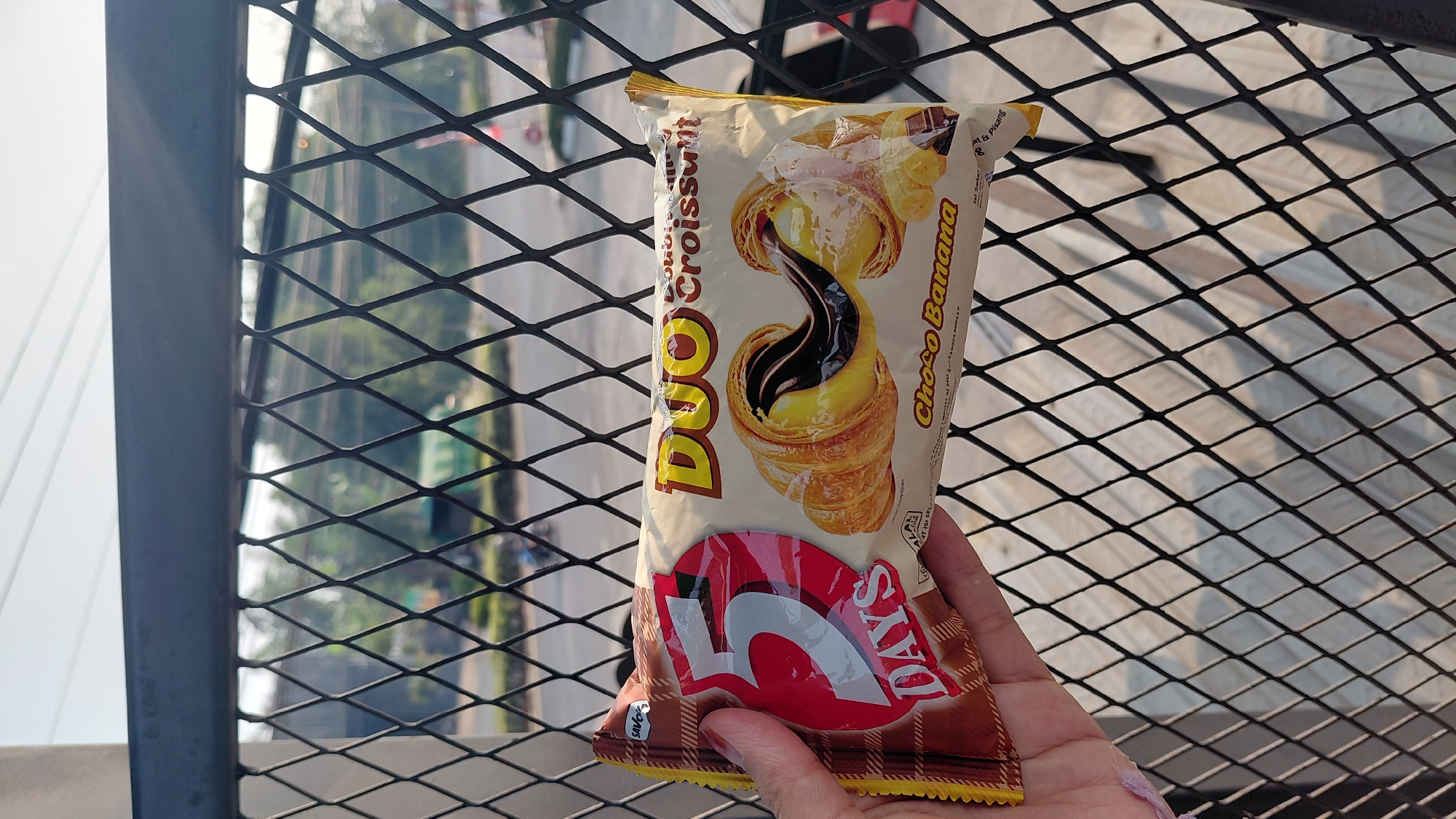 A person is holding a package of "5 Days DUO Croissant Choco Banana" in front of a black metal fence. The package is yellow and brown with an image of two croissants filled with chocolate. The background shows trees and a road. The image appears to have been taken outdoors in daylight.