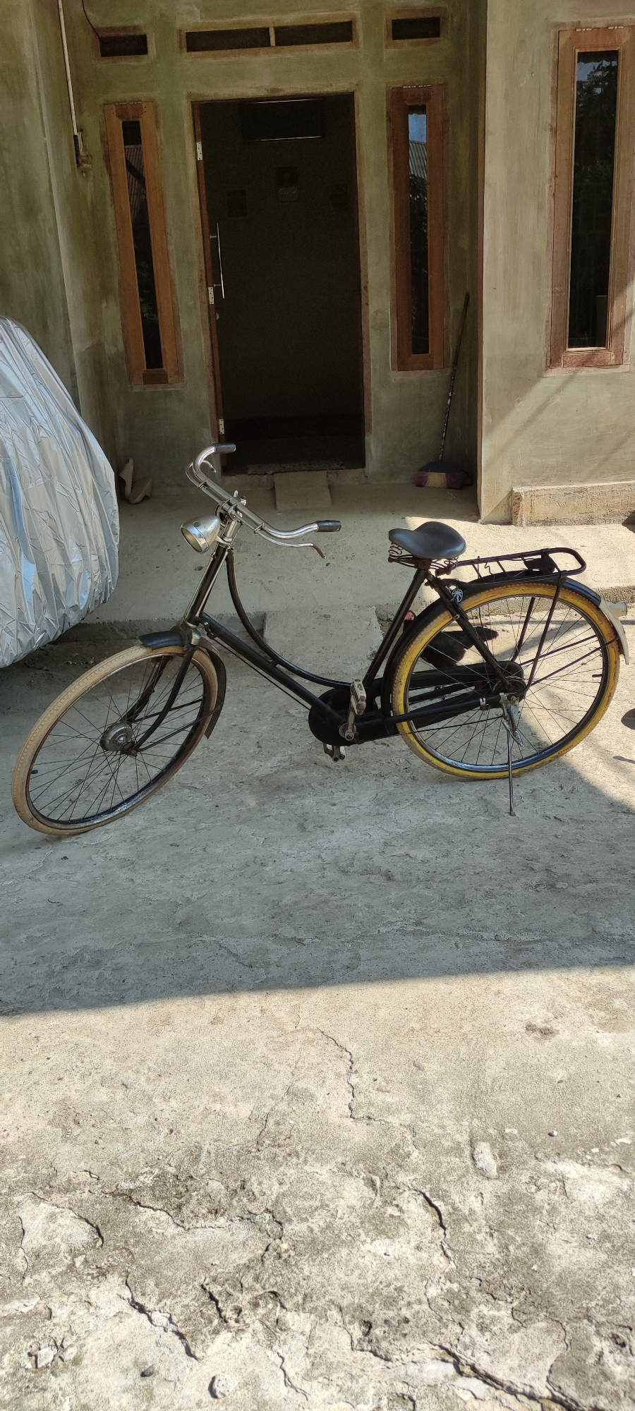 In Payakumbuh, Indonesia, a vintage black bicycle with a silver headlight and a tan tire on the front wheel and a yellow tire on the rear wheel is parked on a concrete surface outside a building. The building has a light-colored, textured facade with a dark-colored open doorway and two narrow, vertical windows with wooden frames. To the left of the bicycle, a silver tarp-covered object is visible. A broom with a colorful head leans against the right side of the building. The ground shows cracks and uneven patches in the concrete.
