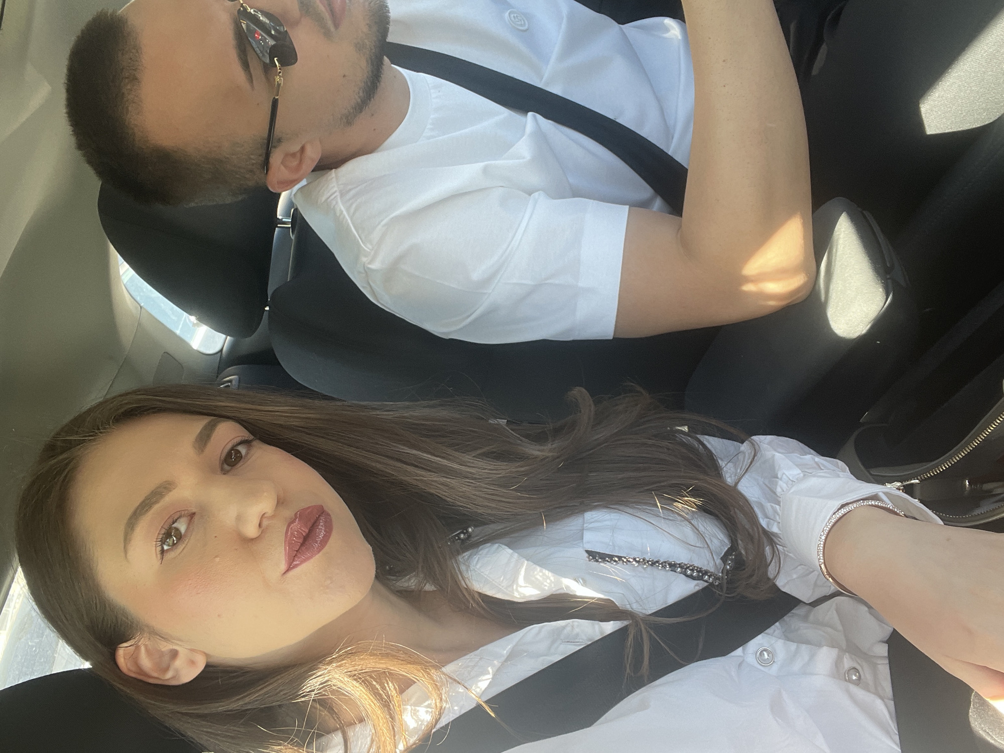 The image shows two people in a car. A woman with long brown hair, wearing a white shirt with silver accents and a bracelet, is in the foreground, seemingly taking a selfie. She is wearing a seatbelt. A man wearing sunglasses and a white shirt is in the background, resting his head on the headrest. He is also wearing a seatbelt. The interior of the car is visible, including the seats and part of a black handbag. The location is likely Sofia, Bulgaria, based on contextual understanding, but there are no elements in the image that can confirm this.