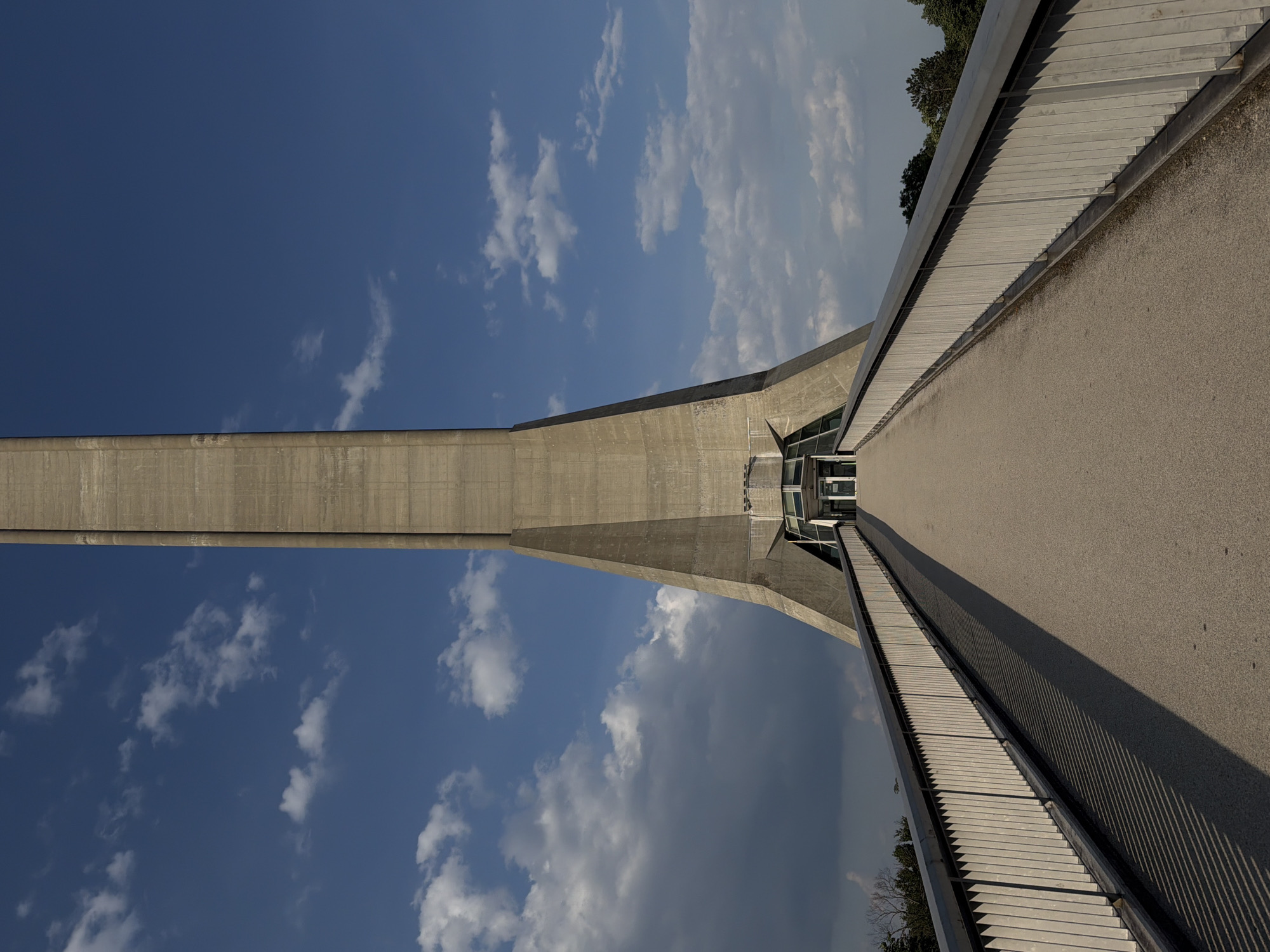 The image shows a low-angle, skewed perspective of the Pylon of Avala, located in Vozdovac Urban Municipality, Serbia. The shot is taken from the walkway leading to the tower's base. The walkway is constructed of concrete and lined with metal railings. The sky, filled with scattered cumulus clouds, is visible behind the pylon. The top of the pylon recedes into the sky. Some trees are visible at the edge of the structure.