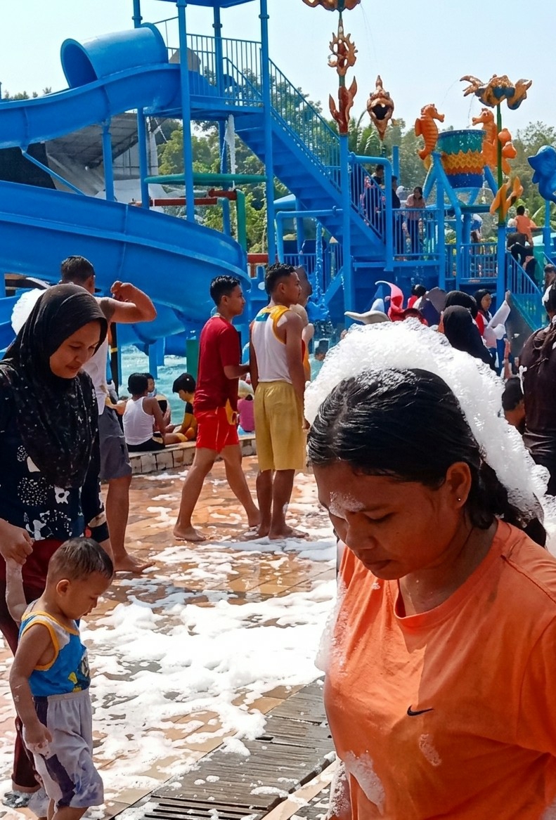 The image, taken in Durin Simbelang, Indonesia, depicts a crowded water park on a sunny day. Dominating the background is a large blue water slide structure with multiple slides, staircases, and decorative aquatic-themed elements. People of various ages are seen on and around the structure. In the foreground, a woman wearing an orange t-shirt is partially covered in white foam and appears to be looking down. Next to her, a young child in a blue and white outfit is also covered in foam. To the left, another woman wearing a dark hijab holds the child's hand. The ground is covered in white foam and wet surfaces. Several other individuals are seen walking or standing in the background, wearing swimwear or casual clothing.