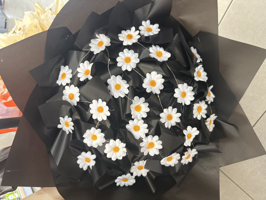 A large floral bouquet featuring numerous artificial daisy-like flowers is displayed indoors in Torbalı, Turkey. Each flower has white petals and a distinct orange center. The flowers are densely arranged and wrapped in crinkled black paper or fabric, which provides a contrasting background. Small, clear lights are subtly integrated and strung throughout the artificial flowers. The bouquet rests on a light-colored tiled floor with visible grout lines. In the soft-focus background, fragmented views of other floral arrangements or wrapping materials, including yellow and orange elements, are discernible, suggesting a retail setting, likely a flower shop or gift store. No individuals or active interactions are visible.
