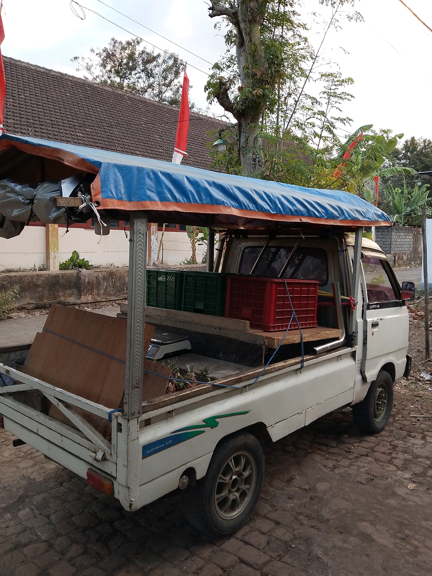 A white flatbed pickup truck, identified as a "Carry 1.0" on its side, is parked on a cobblestone street in Karangsari, Indonesia. The truck is covered with a blue tarp with an orange trim, supported by metal poles. The truck bed is loaded with several items, including large wooden planks, two green plastic crates, and a red plastic crate. A digital scale is visible in the truck bed, alongside some pineapples. The background shows a traditional tiled roof house with red and white Indonesian flags displayed. Trees and vegetation are abundant, indicating a semi-rural or suburban setting. The sky is overcast, suggesting it might be late afternoon or early morning. There are no people visible in the image.