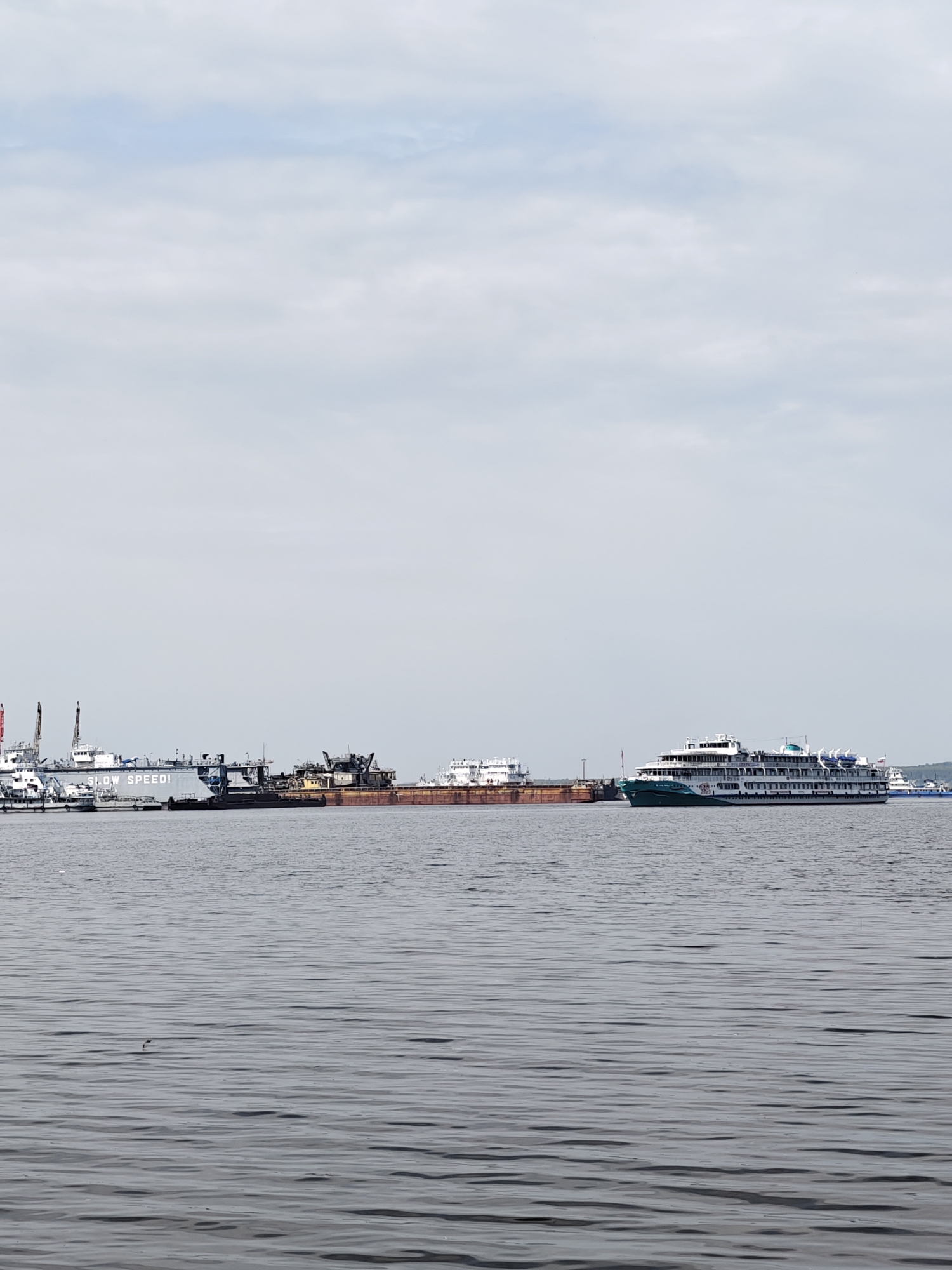 The image depicts the Kama River in Chaikovsky, Russia. The foreground is the river's gray, rippled surface. In the middle ground, several vessels are docked along the riverbank. From left to right, there is a dredger with a sign that reads "SLOW SPEED!", a rusty barge, and a river cruise ship with green and white accents. Further in the distance, a smaller boat is visible. The background is dominated by a cloudy, gray sky.