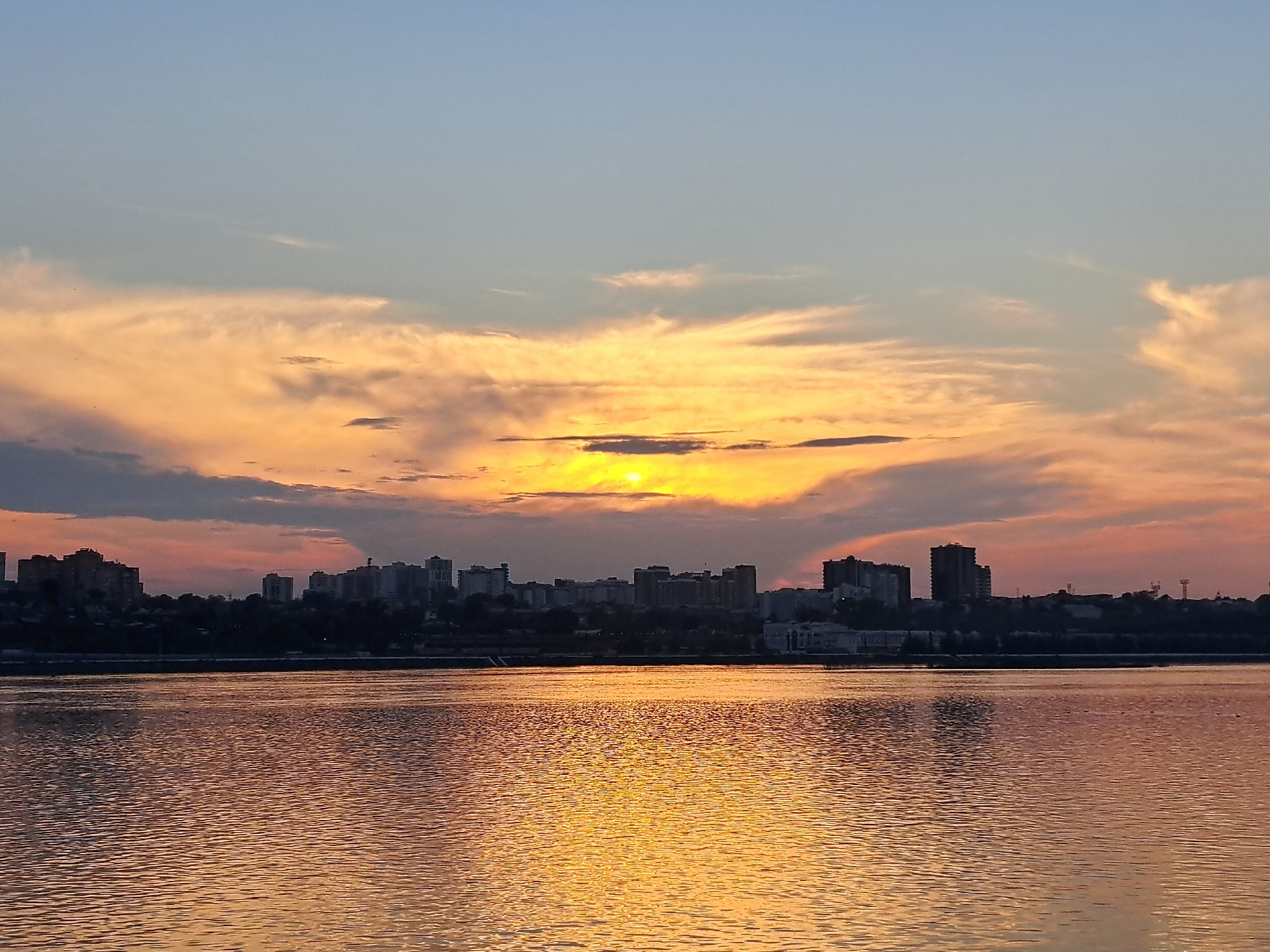 The image captures a sunset over the Angara River in Irkutsk, Russia. The sky displays a gradient of soft blues and oranges, with wispy clouds catching the last rays of the sun. The sun itself is partially obscured by clouds, casting a warm, golden glow onto the water. In the distance, the silhouette of the Irkutsk cityscape is visible, featuring a line of buildings against the horizon. The river's surface is rippled, reflecting the vibrant colors of the sky and the city lights. The scene conveys a sense of calm and tranquility.