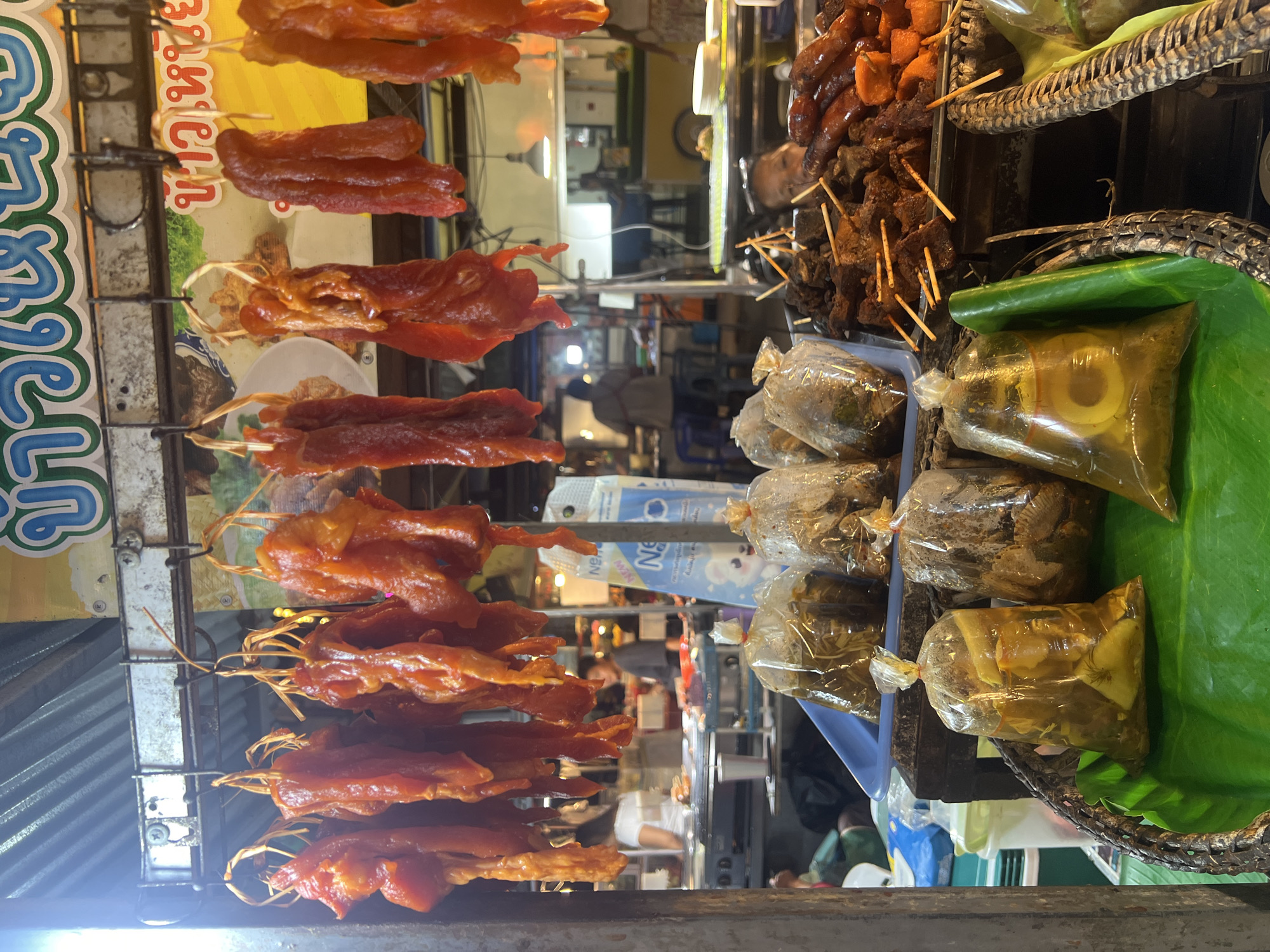 The image depicts a street food stall in Koh Phangan, Thailand. The foreground features a woven basket filled with various food items, including plastic bags containing pickled or preserved foods, and a stack of grilled meat skewers. The meats on skewers vary in color and texture, with some appearing to be dark brown and others a reddish-orange.
Behind the basket, several rows of marinated, red meat strips are hanging from metal hooks. These strips are various shades of red and orange, suggesting they are being cured or dried. They are suspended by thin twine.
In the mid-ground and background, the stall appears to be bustling. People can be seen behind the food displays, some of whom are likely vendors or customers. There are food preparation surfaces, containers, and other stalls visible in the distance, illuminated by overhead lighting. A blue and white container, possibly for drinks or ice, is also visible. The overall environment suggests a lively outdoor market or street food area.